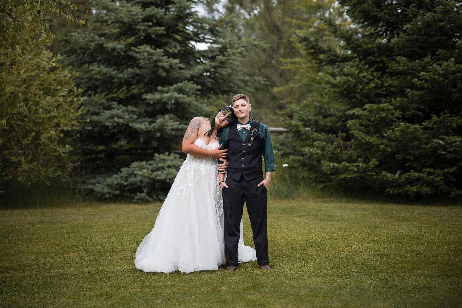 A couple embraces on their wedding day while standing on a lush lawn surrounded by evergreen trees.