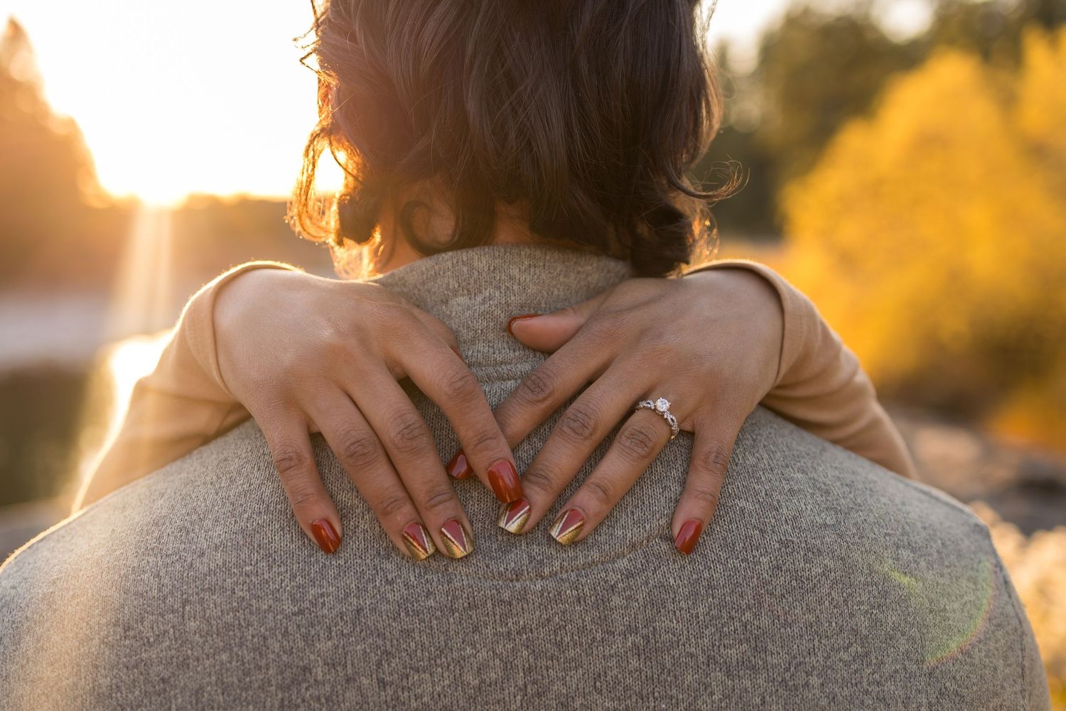 Close-up view of hands embracing from behind during golden sunset light with visible engagement ring.