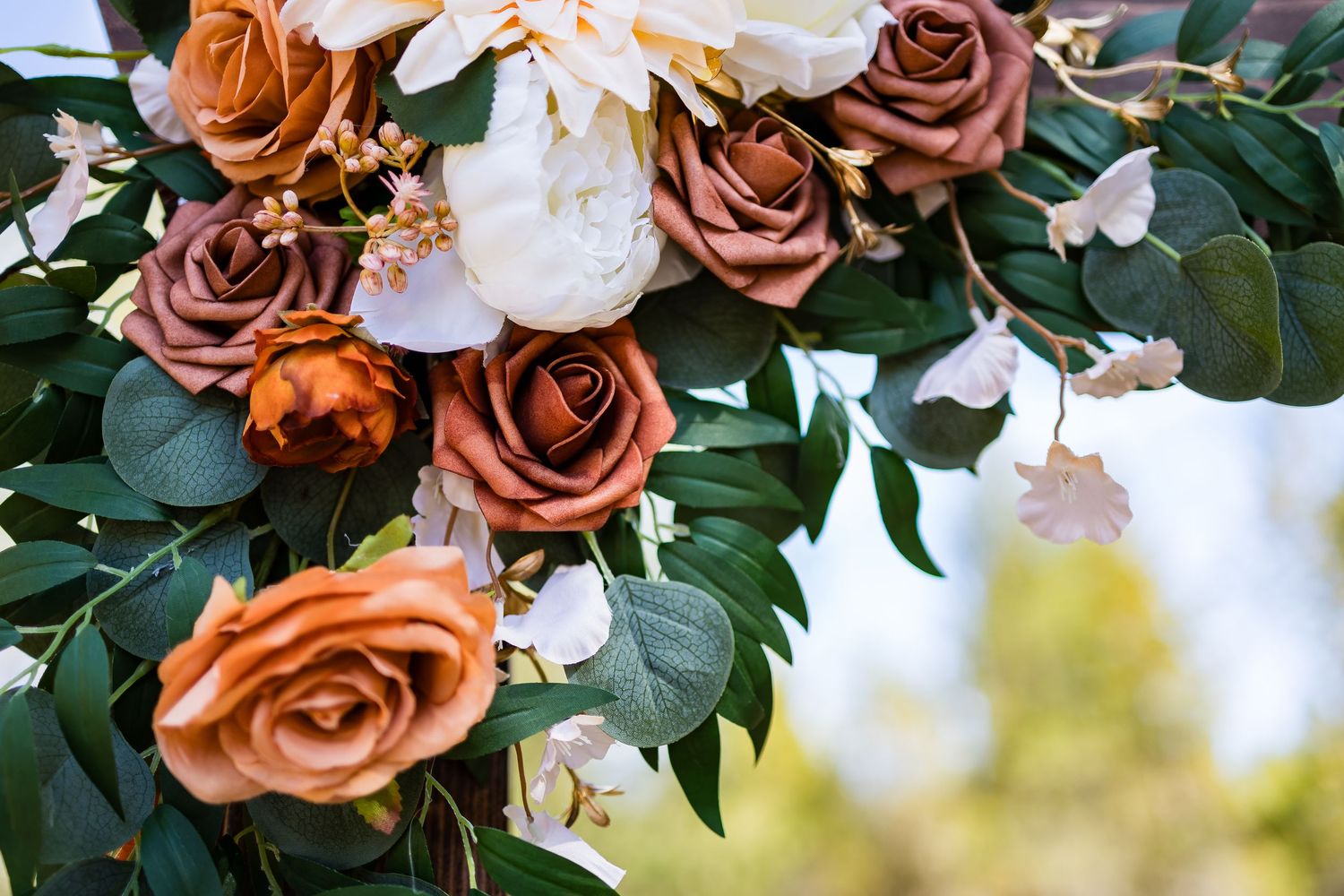 A floral arrangement with rust-colored roses and white peonies decorated with green eucalyptus leaves against a blurred background.