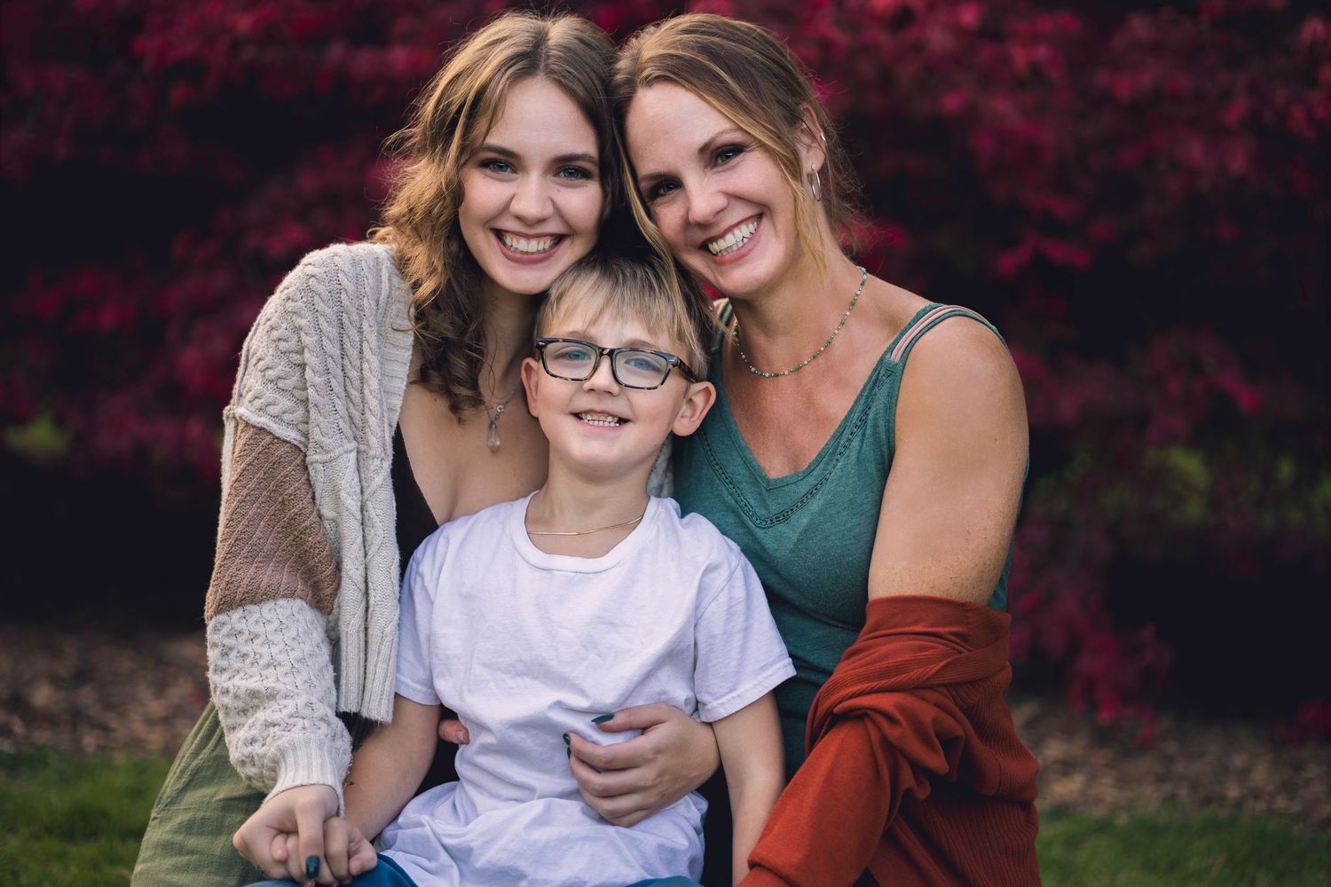 Two adults pose with a child wearing glasses against a background of deep red autumn foliage.