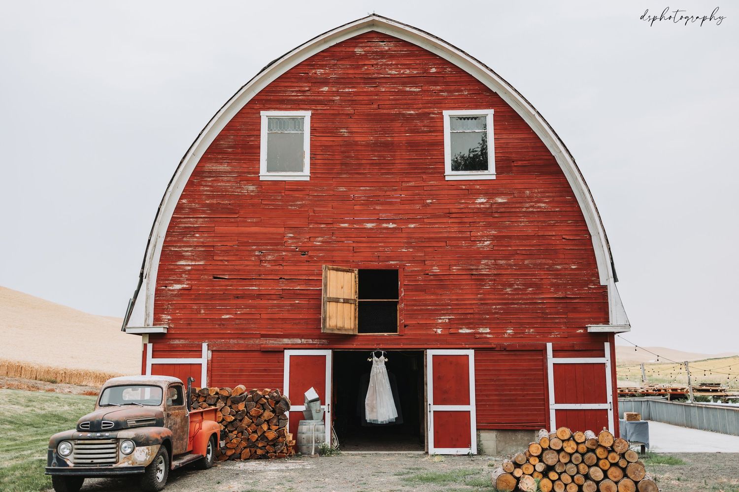 Rustic red barn with vintage truck and stacked firewood displays a white wedding dress hanging in the doorway.