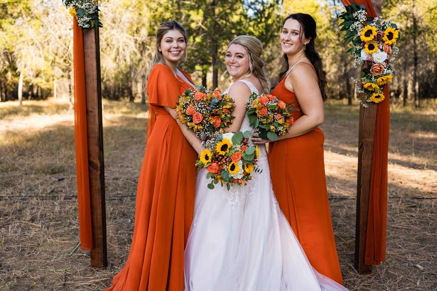 Autumn outdoor wedding party poses with bright orange dresses and vibrant sunflower bouquets in wooded setting.
