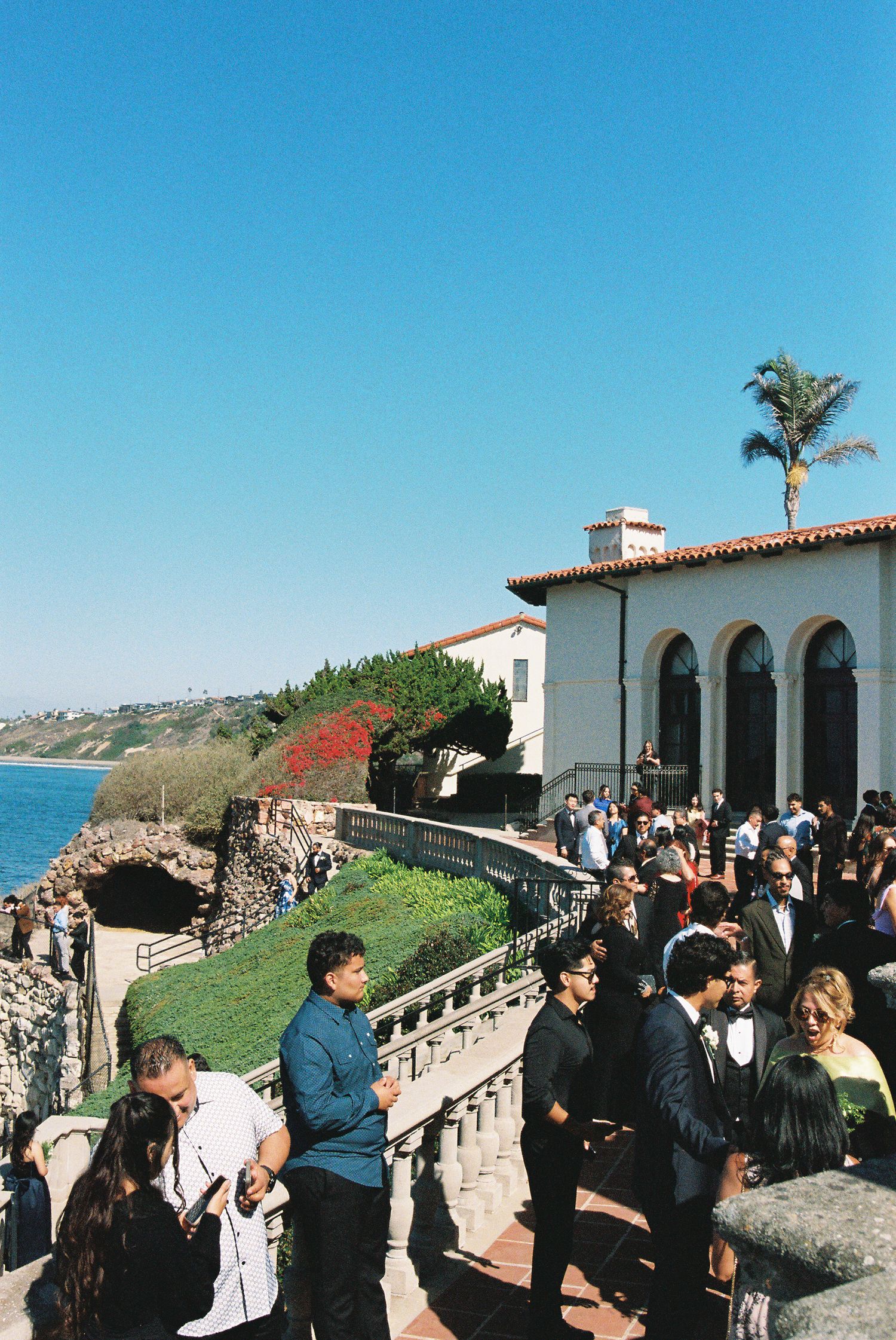 Panoramic view of people gathering at a coastal Spanish-style building with arches and ocean views on a sunny day.