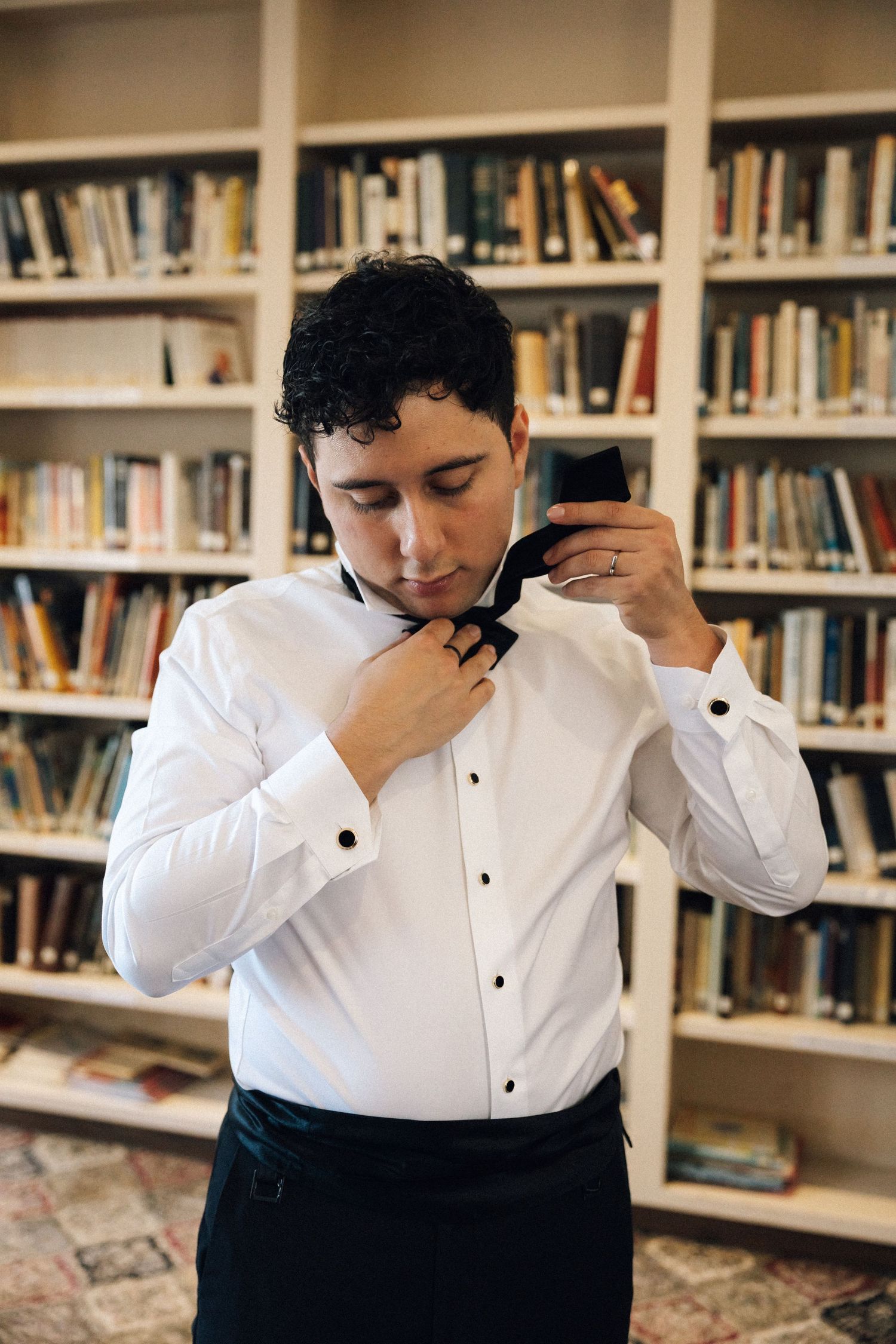 Person in white dress shirt and black pants stands thoughtfully in library with bookshelves in background.