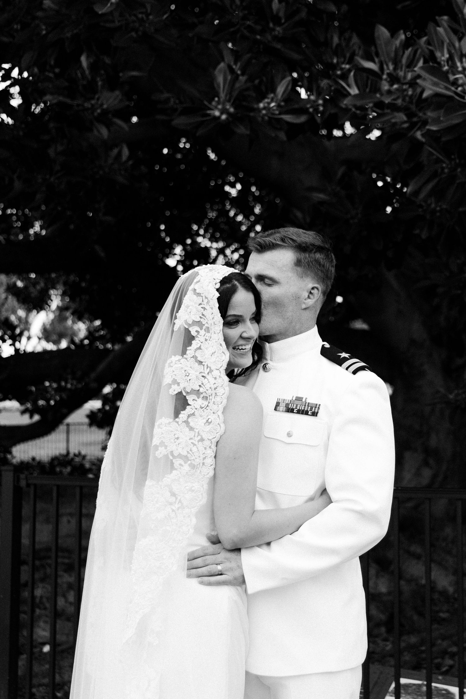 Black and white photo of a bride in lace veil and groom in white military uniform sharing a tender kiss outdoors.