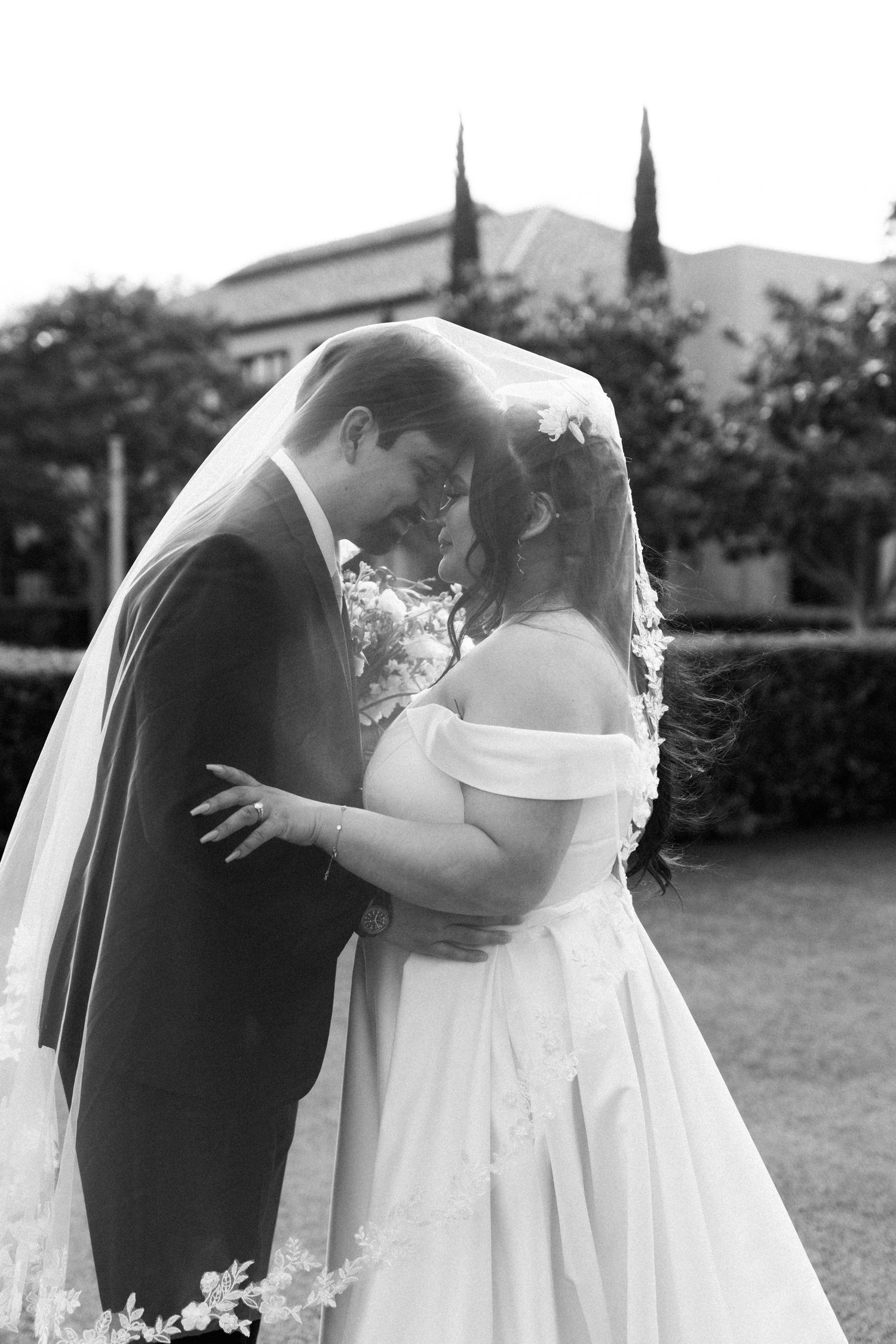 A romantic black and white wedding photo shows a couple embracing outdoors with the bride in an off-shoulder gown and flowing veil.