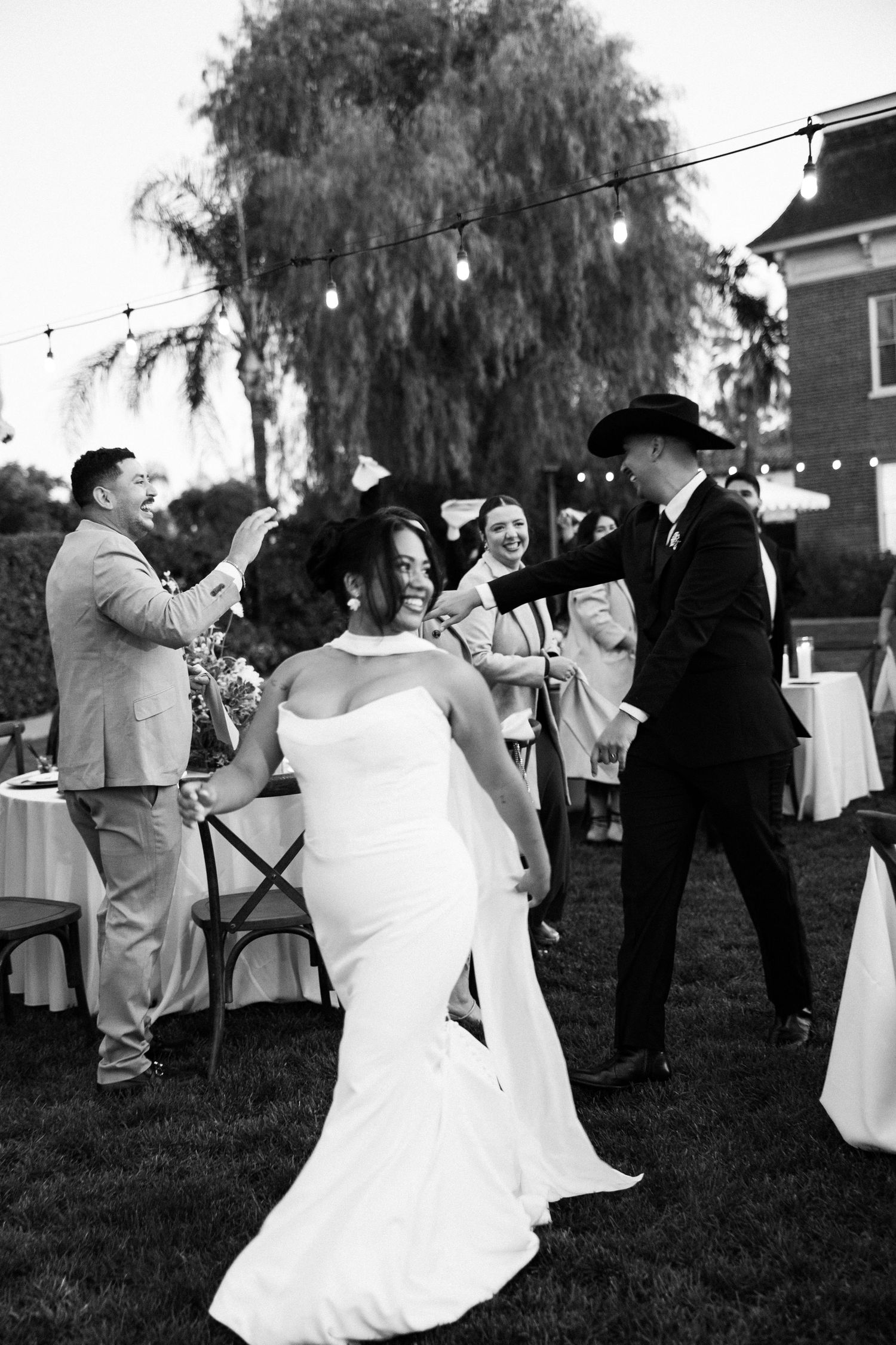 Wedding couple dances together at outdoor reception under string lights and willow tree in black and white photo.