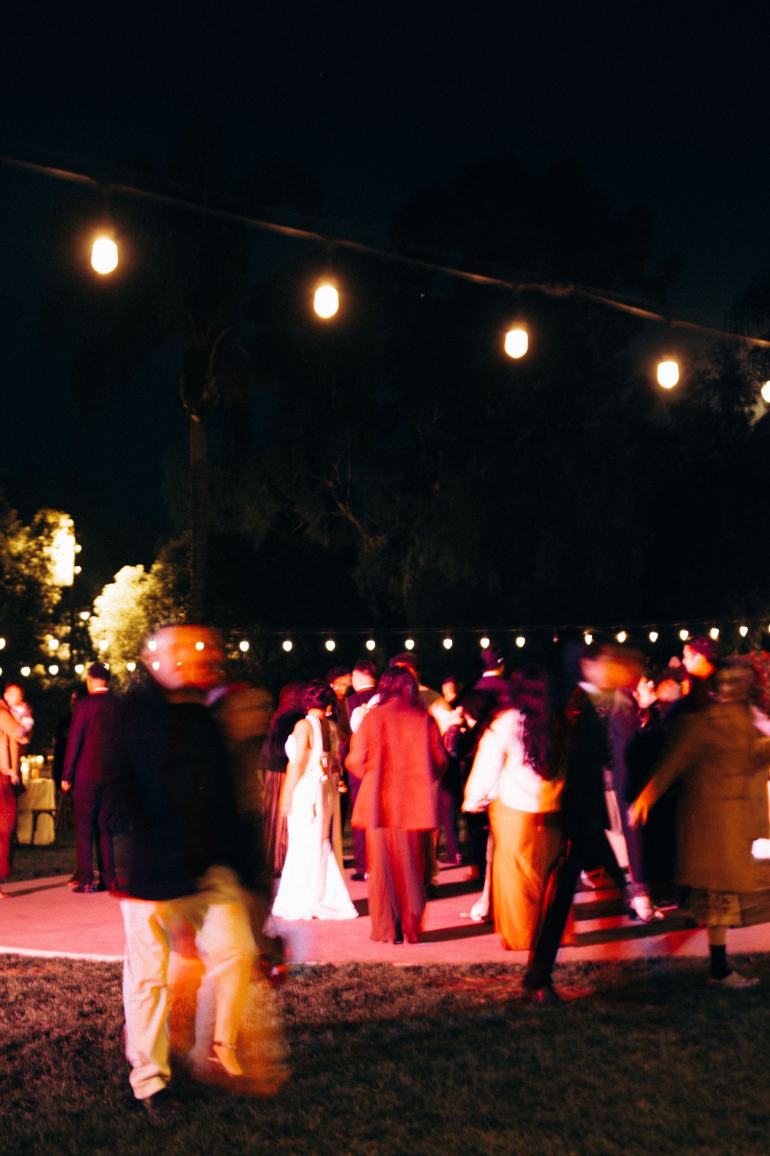Guests gather at an outdoor evening event under glowing string lights with pink and purple ambient lighting.