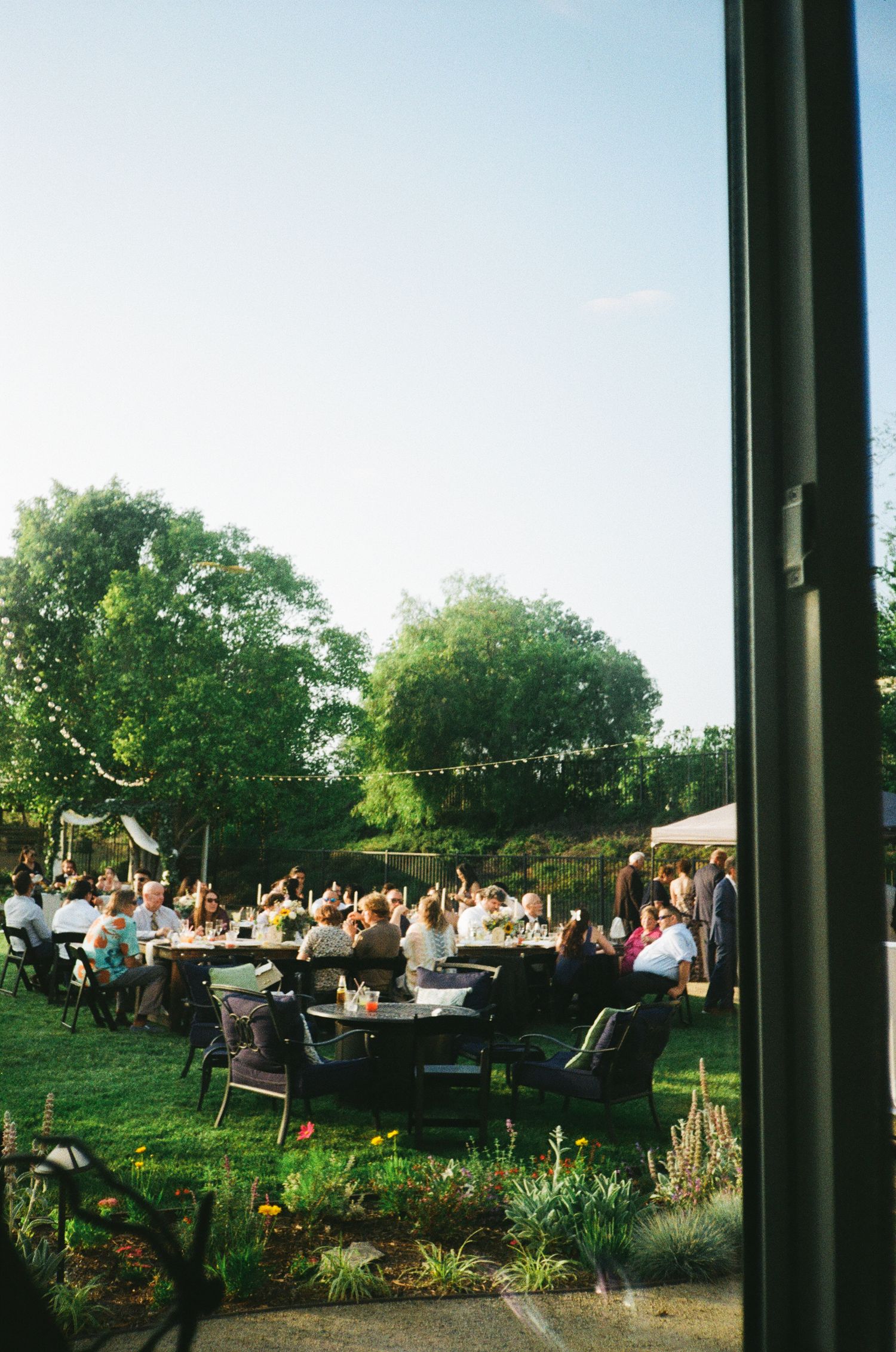 Guests gather at outdoor tables for a garden party reception under string lights and trees at dusk.