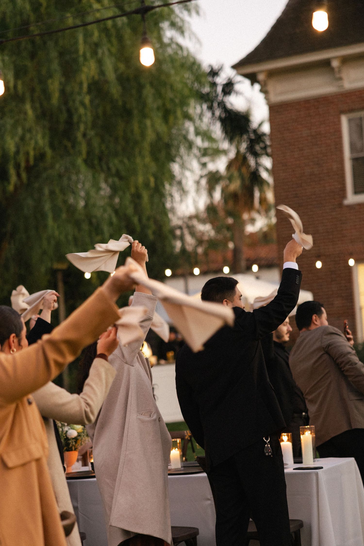 Wedding guests dance and raise their arms in celebration at an outdoor evening reception with string lights overhead.