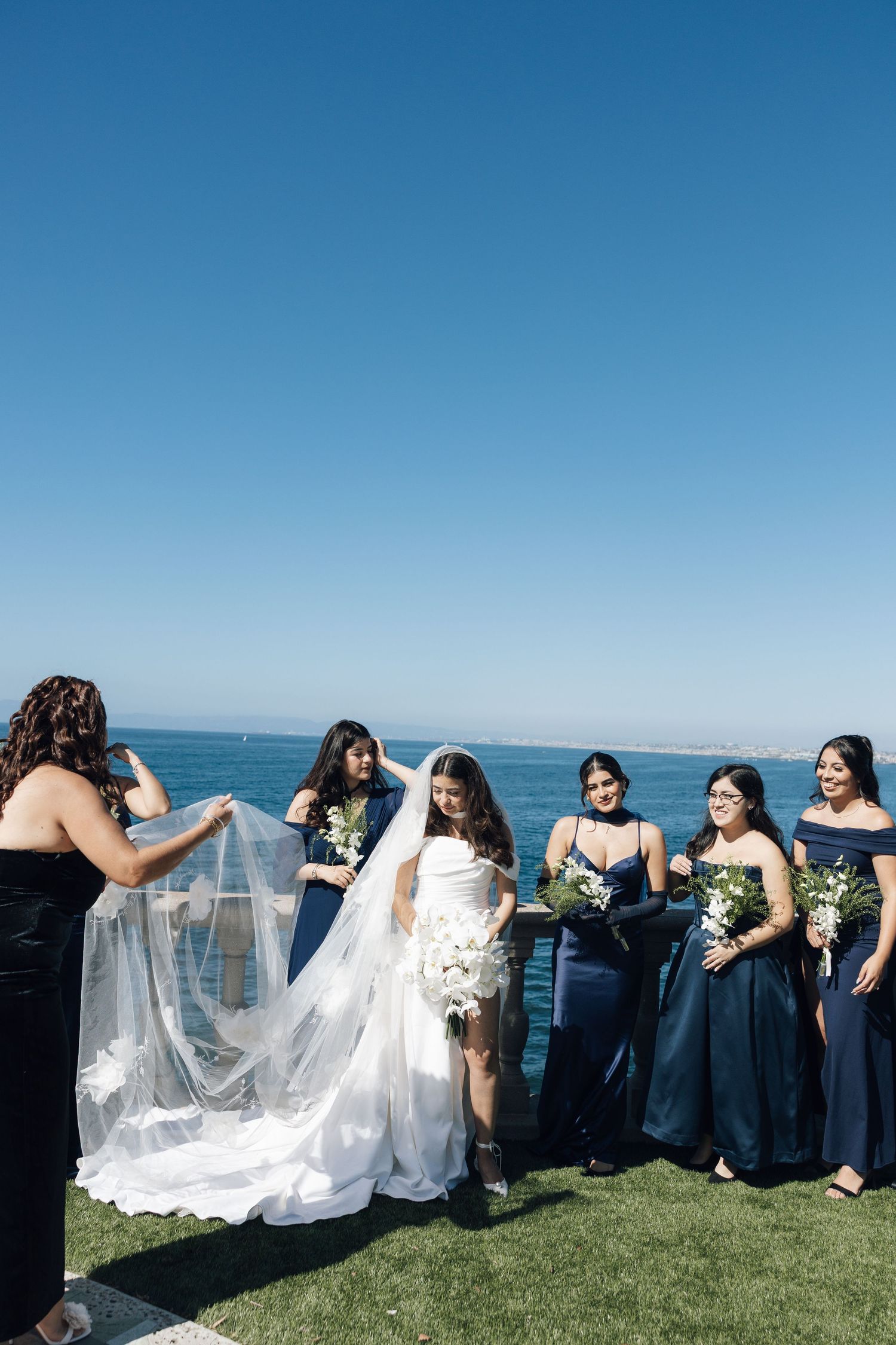 Bridesmaids in navy blue dresses help arrange the bride's flowing white veil on a grassy oceanside cliff.