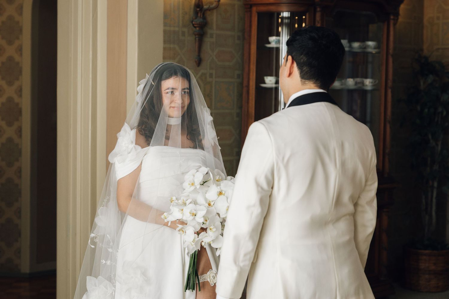 A bride in a white wedding dress and veil stands in an elegant hallway with ornate architectural details.