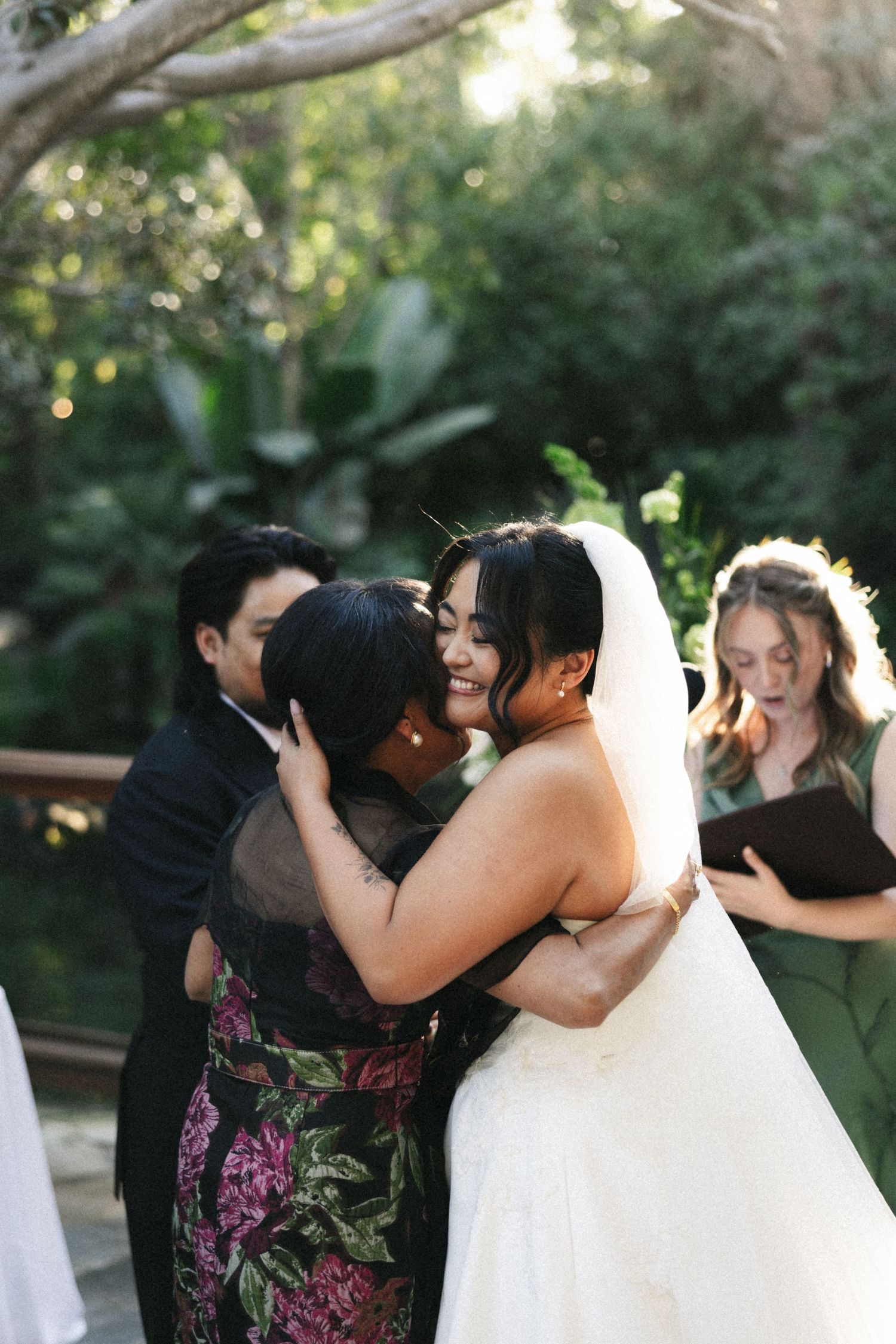 Bride in white gown and veil shares a warm hug with a guest in floral dress during an outdoor wedding ceremony.
