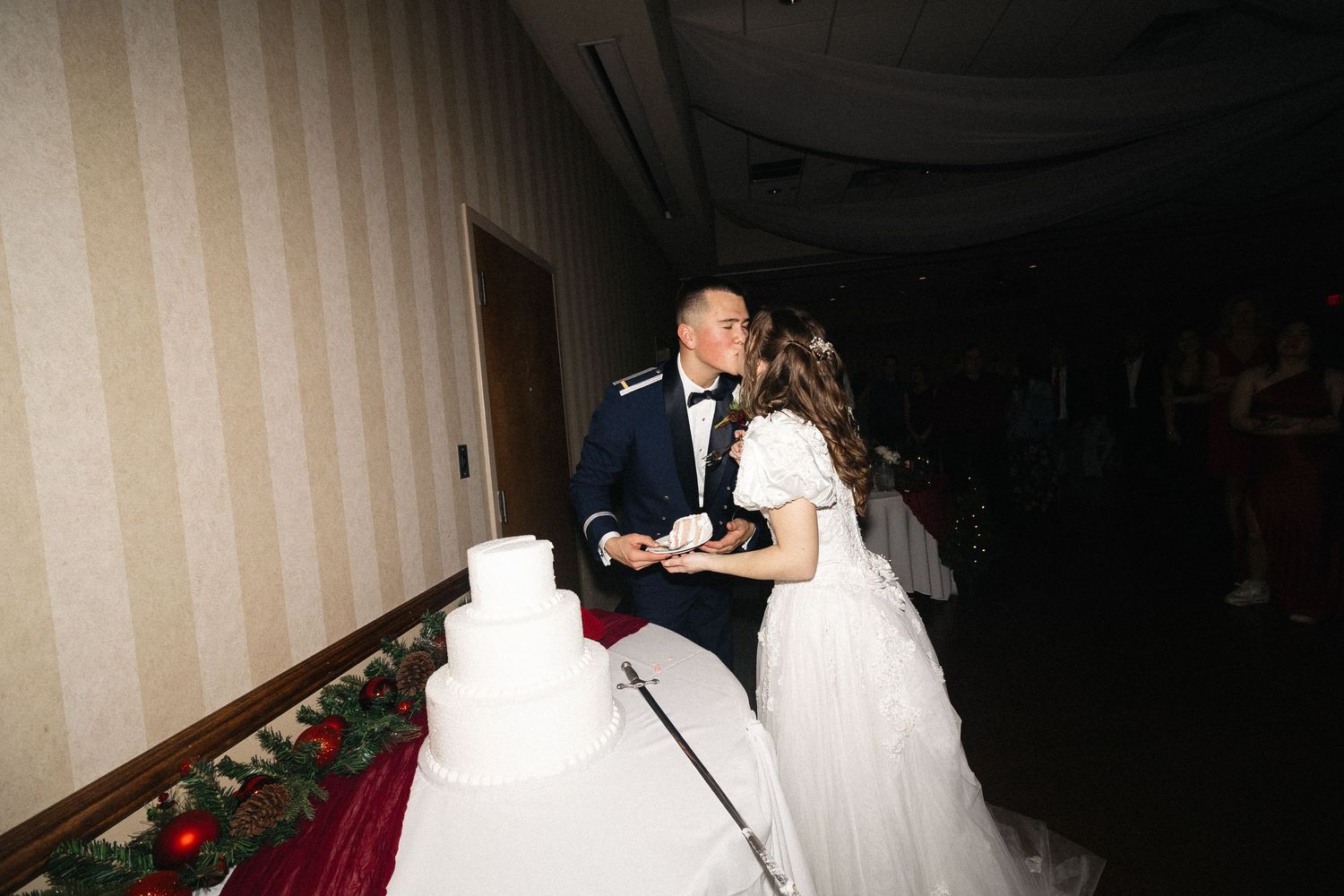 A couple in white wedding attire cuts their tiered white wedding cake together in a dimly lit reception venue. Documentary/ photojournalist