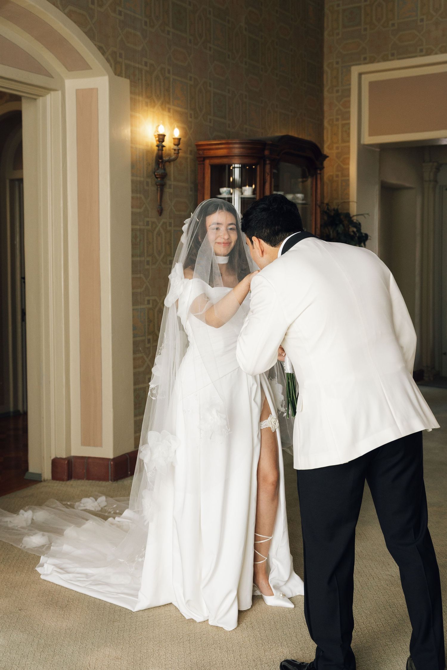 A wedding photographer adjusts the bride's veil in an elegant hallway with warm lighting and ornate walls.