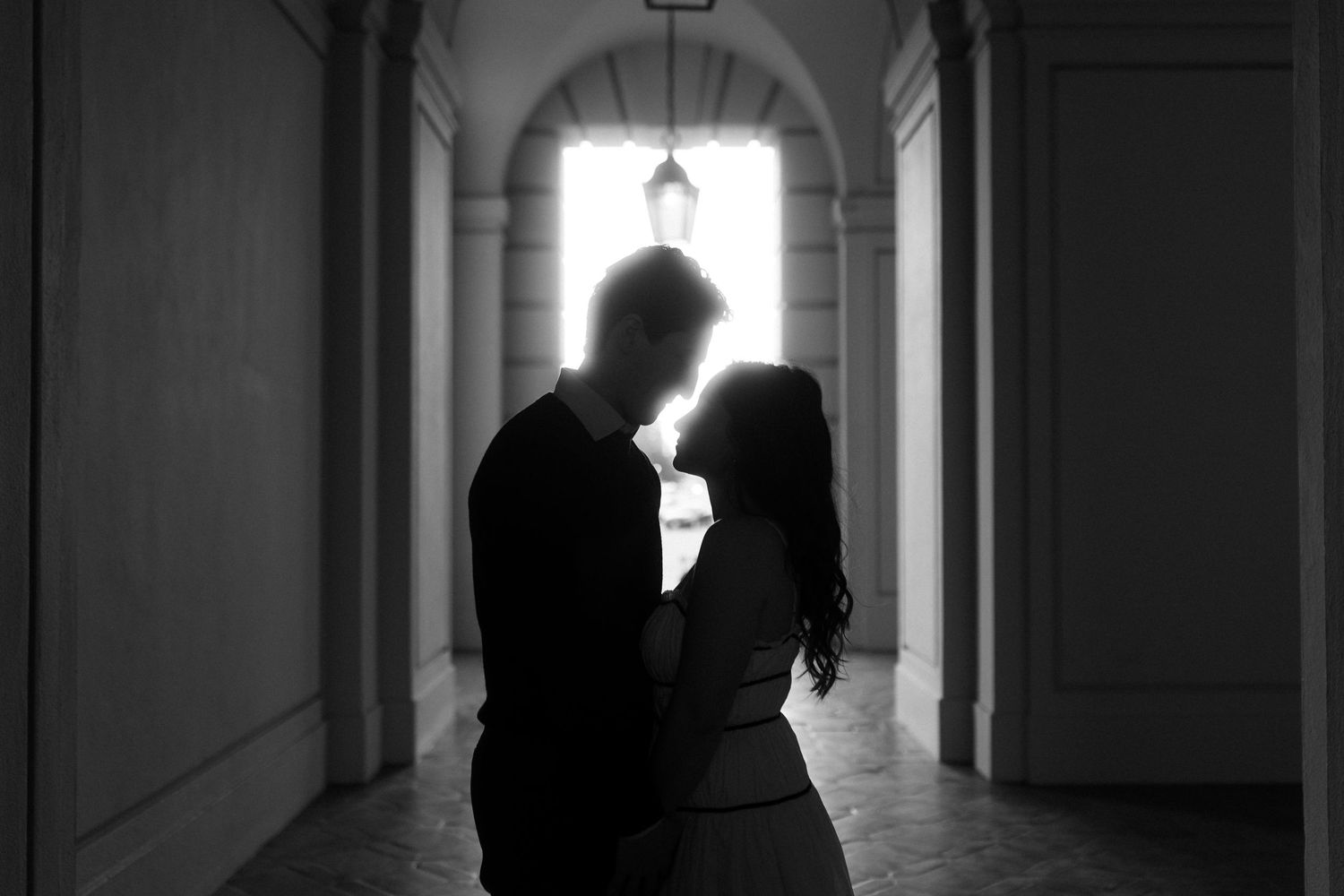 A silhouetted couple stands in a romantic embrace within an arched hallway backlit by bright natural light. Documentary photographer