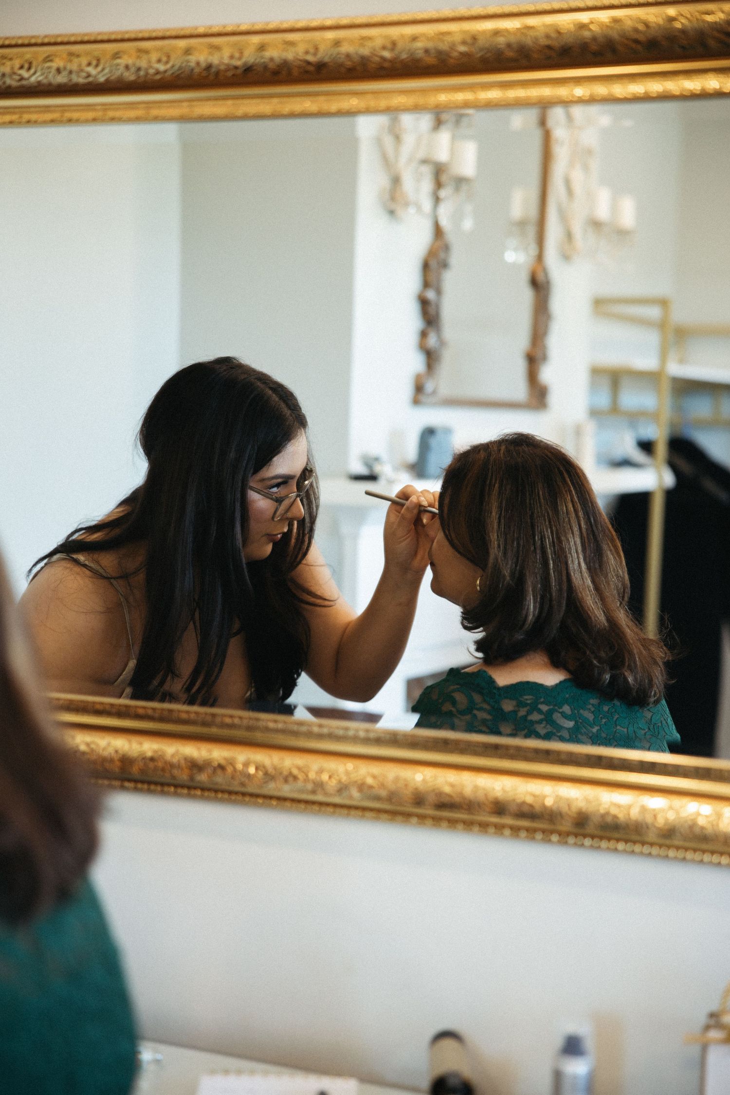 A person applies makeup while looking in an ornate gold-framed mirror.