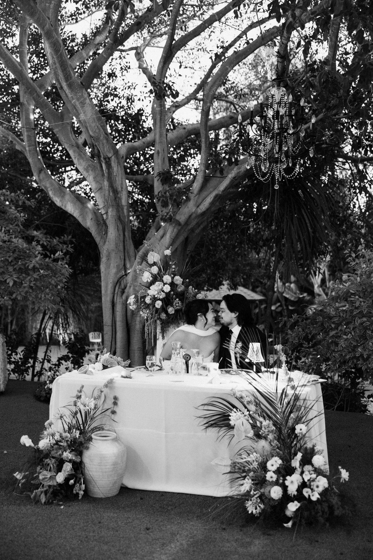 Black and white photo of a couple kissing at a floral-decorated sweetheart table outdoors under a large tree.