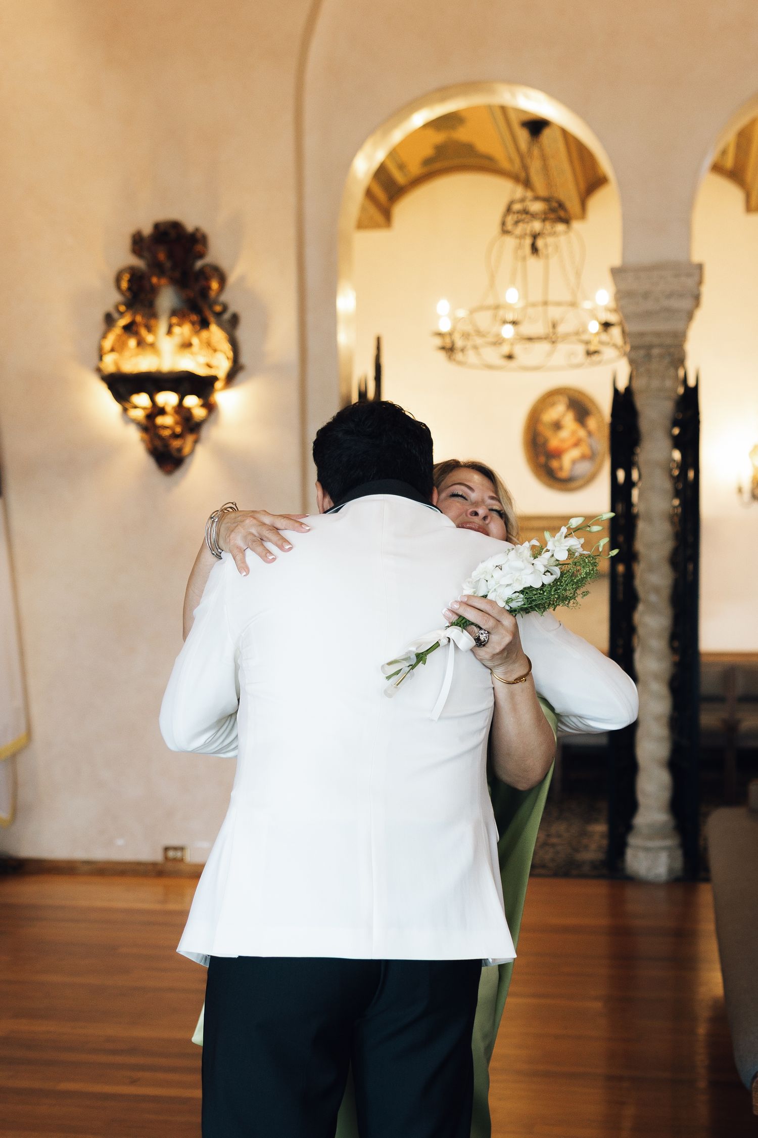 Two people share an intimate embrace during a wedding ceremony in an elegant venue with arched doorways and ornate lighting.