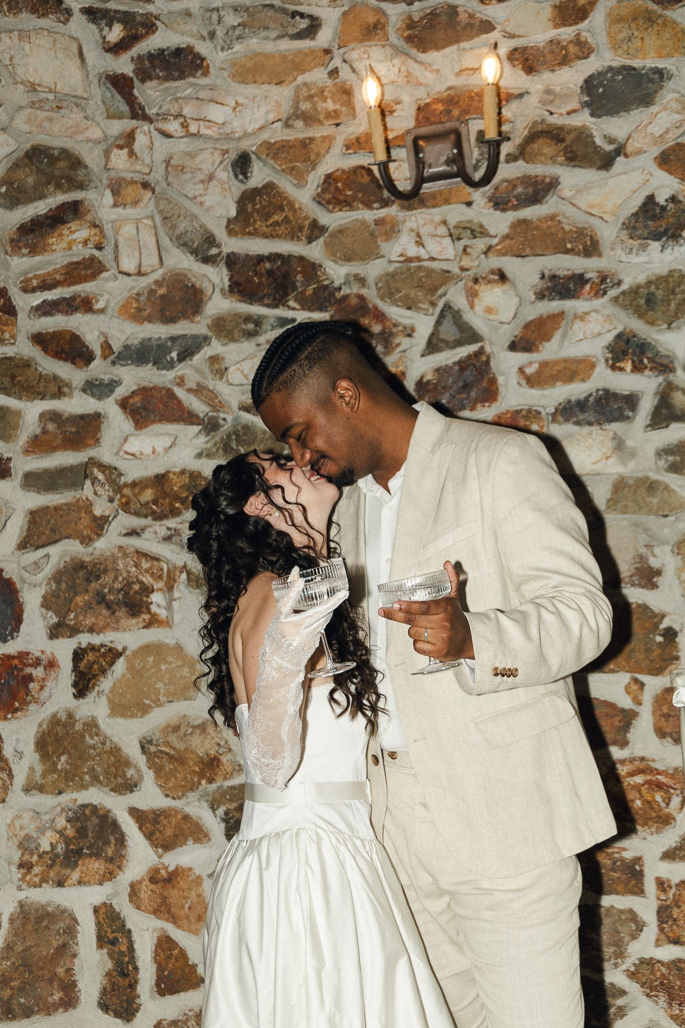 A couple in wedding attire share an intimate moment against a rustic stone wall background. Micro wedding, intentional celebrations