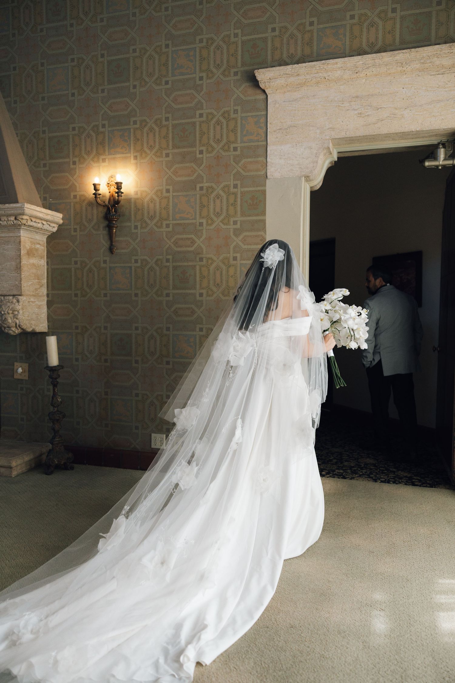 A long flowing white wedding veil trails behind a bride in a classic wedding gown against an ornate wallpapered interior.