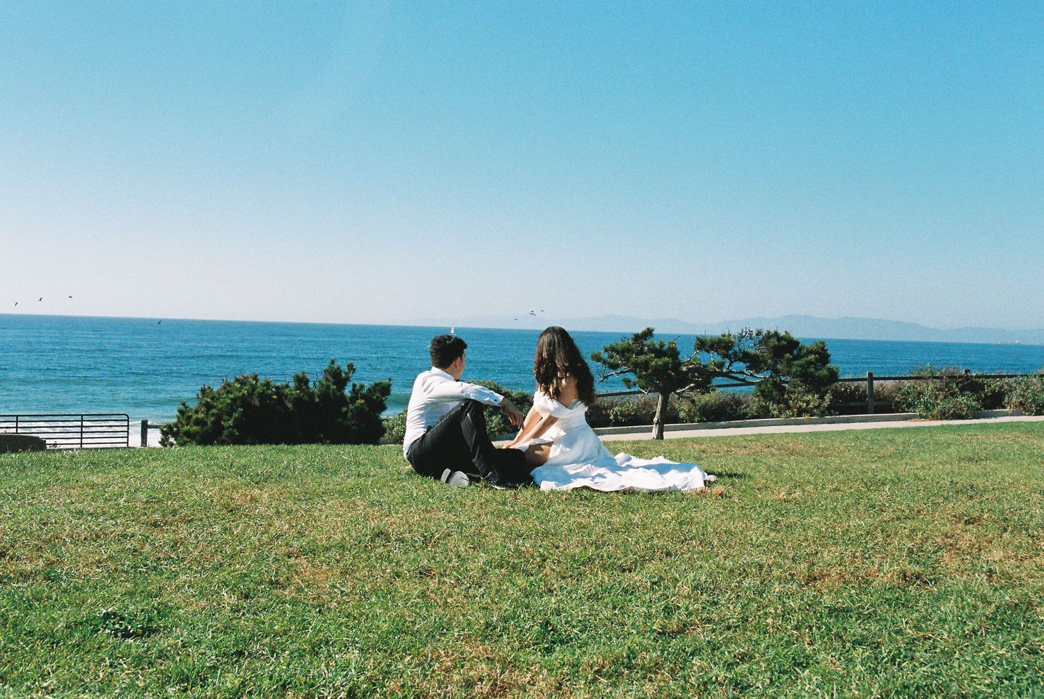 Two people enjoy a scenic picnic on a grassy hillside overlooking the ocean on a sunny day.