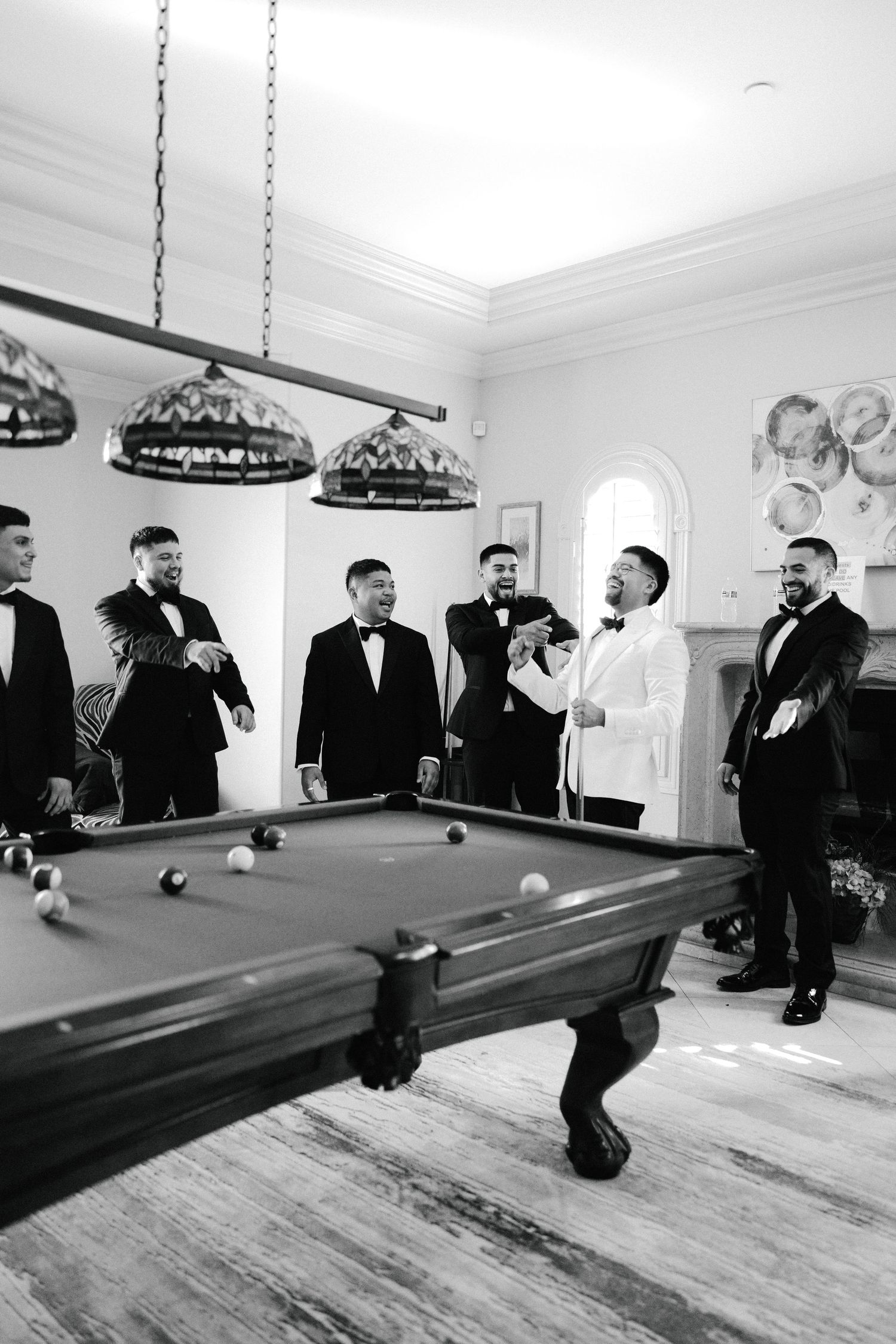 Groom in white tuxedo celebrates with groomsmen in black tuxedos around a pool table in elegant billiard room.