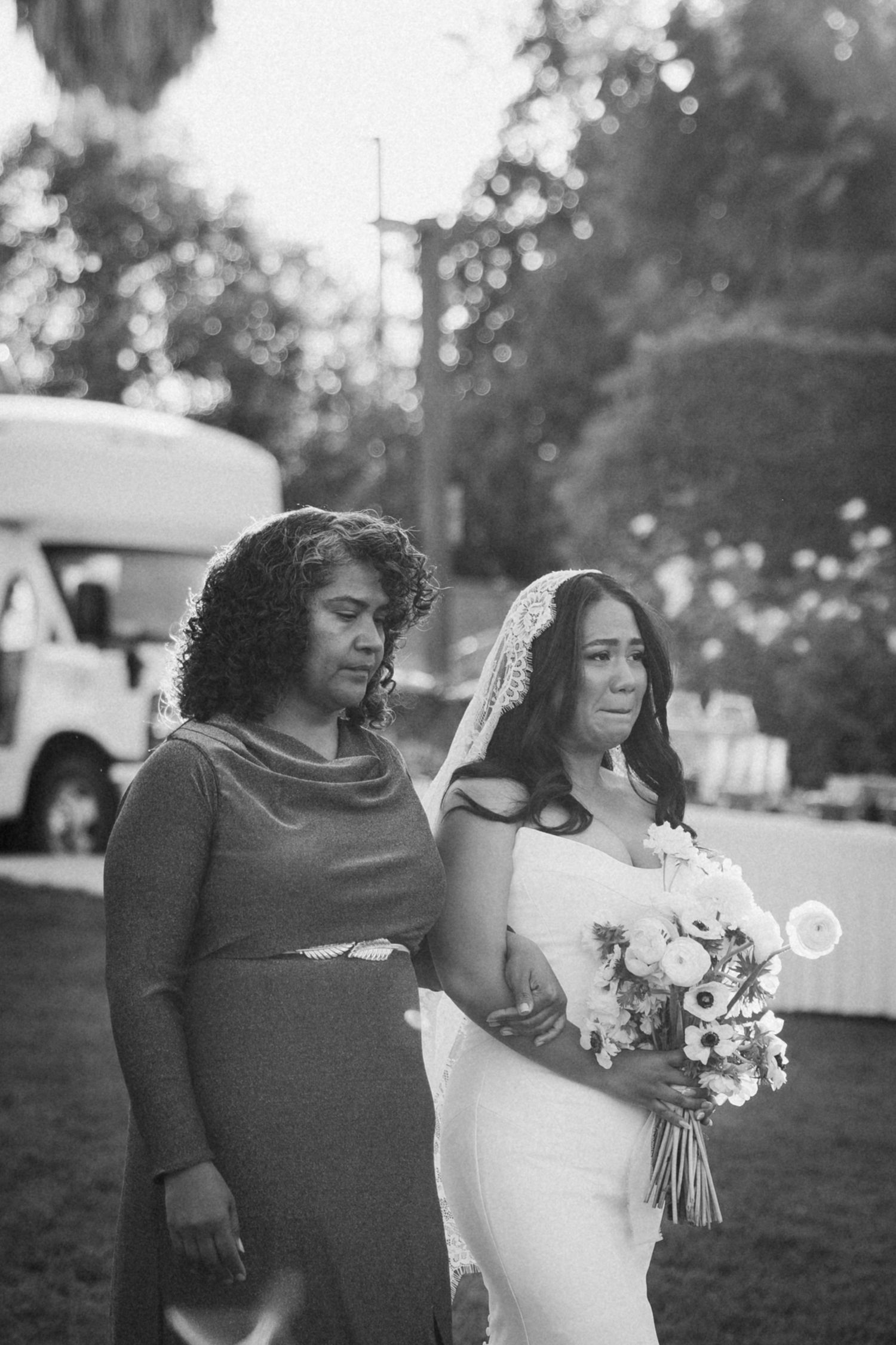 Black and white photo of a bride in a veil holding a bouquet walking with a woman in a dress at an outdoor wedding.