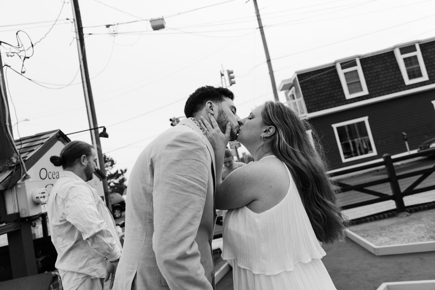 Couple sharing a romantic kiss outdoors in black and white wedding photo near a coastal building.