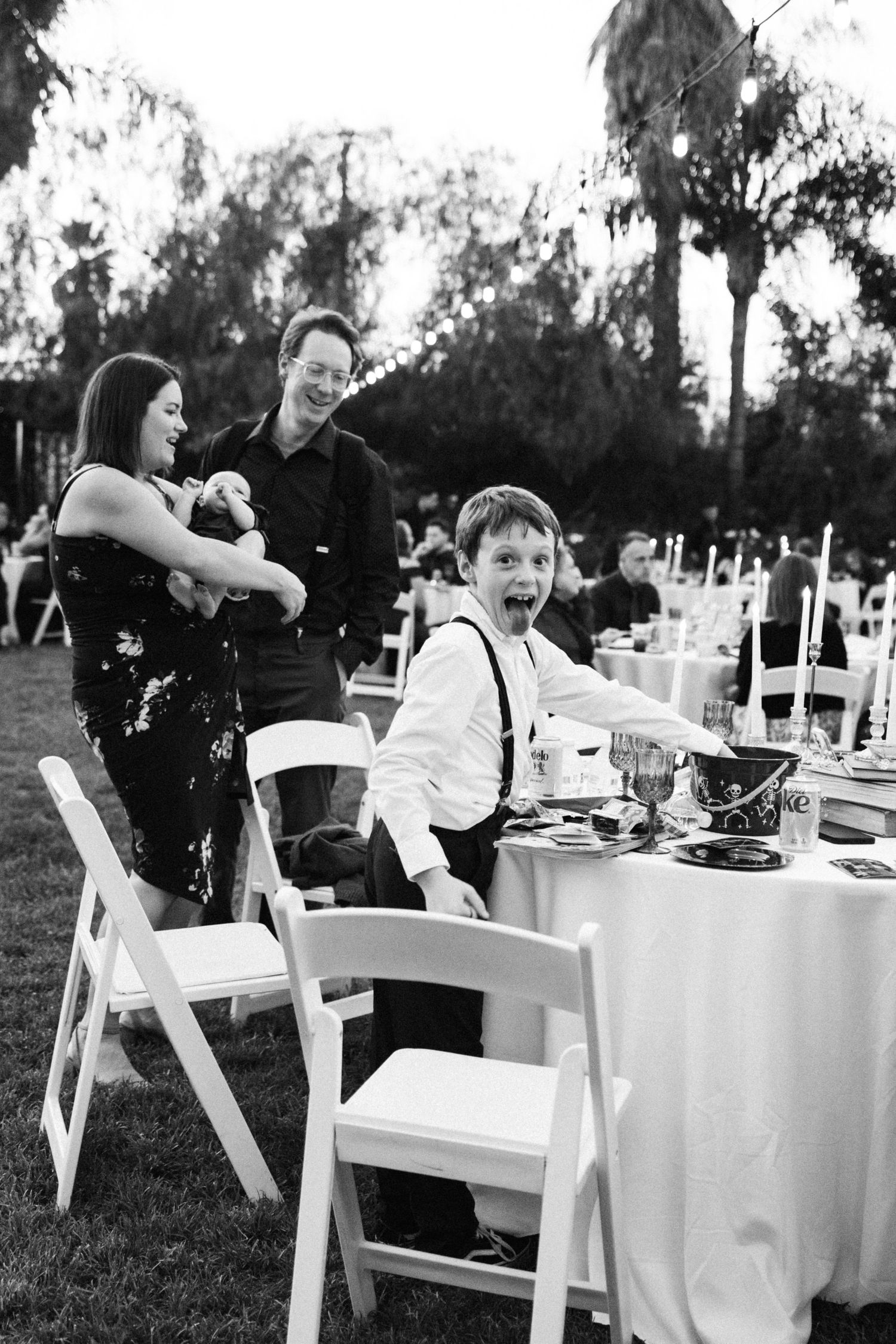 Black and white candid photo of people at an outdoor wedding reception with white tables and chairs on a lawn. Micro wedding, intentional ce