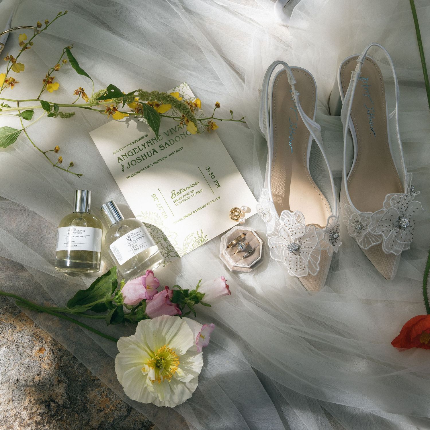 Elegant bridal flat lay with white heels, perfume bottles, wedding rings, lace garter, flowers, and stationery on tulle fabric.