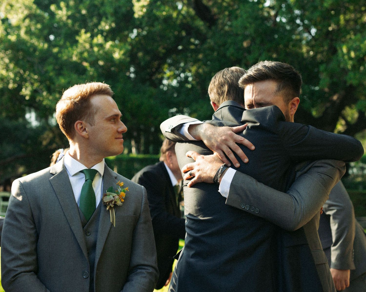 A group of men in formal suits share an emotional embrace and celebrate outdoors during a wedding ceremony. Documentary/ photojournalist pho