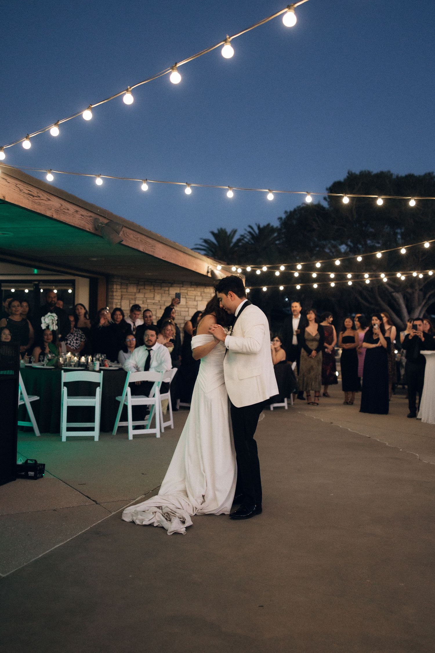 A couple shares their first dance under romantic string lights at an outdoor evening wedding reception.