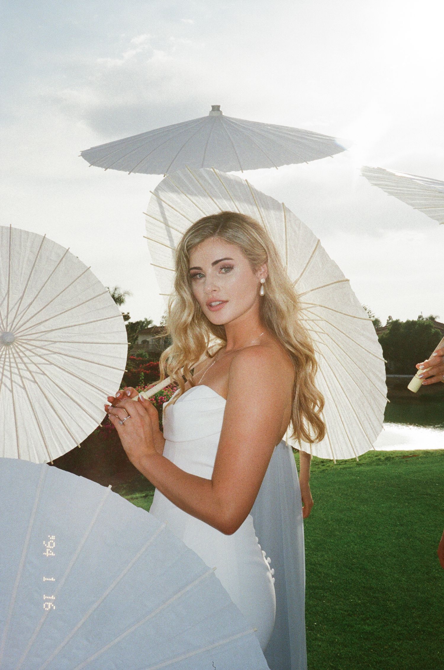 A bride in a white strapless gown poses outdoors holding a white paper parasol against a cloudy sky background.