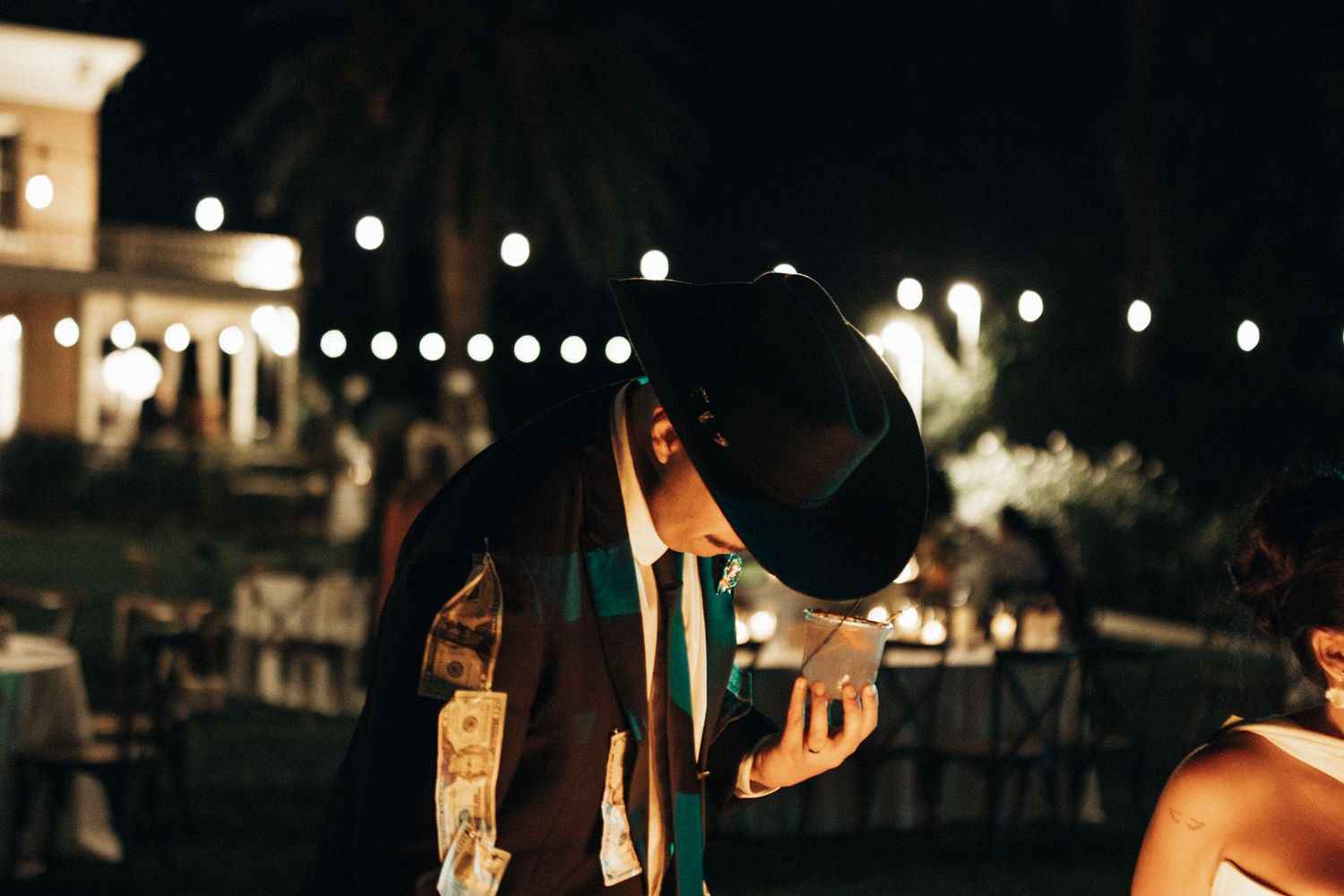 A person in a black hat and suit stands at an outdoor evening event with twinkling lights in the background.