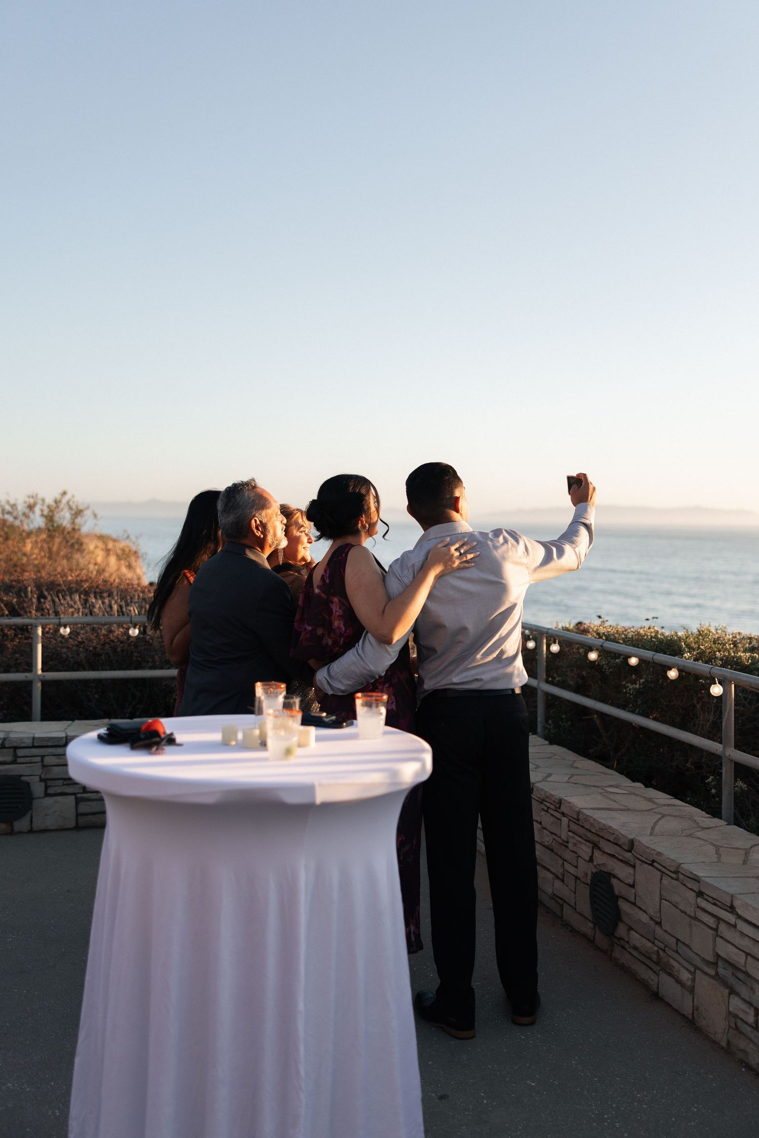 Guests gather around white cocktail tables at an outdoor oceanfront event during sunset.