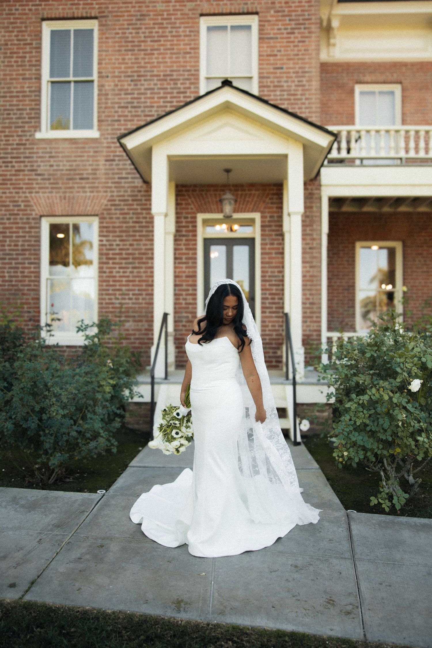 A bride in a flowing white wedding gown sits gracefully on stone steps in front of a brick building.