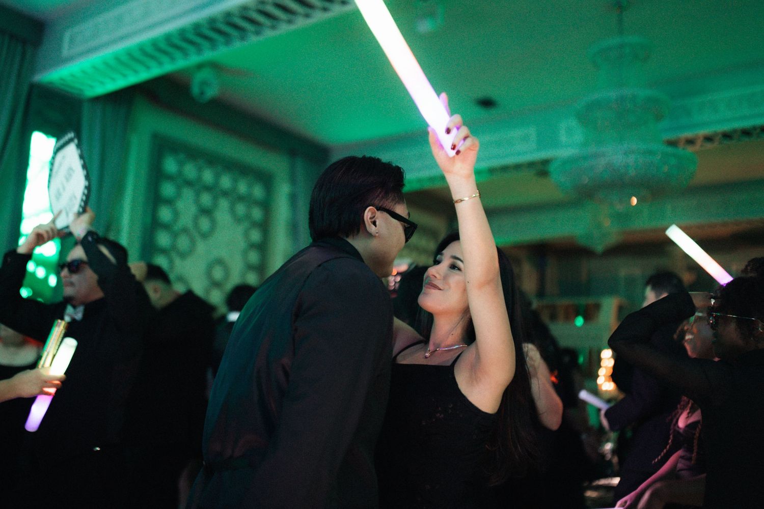 People dance and celebrate under green lighting at a party venue with glowing light sabers raised in the air. Micro wedding, intentional