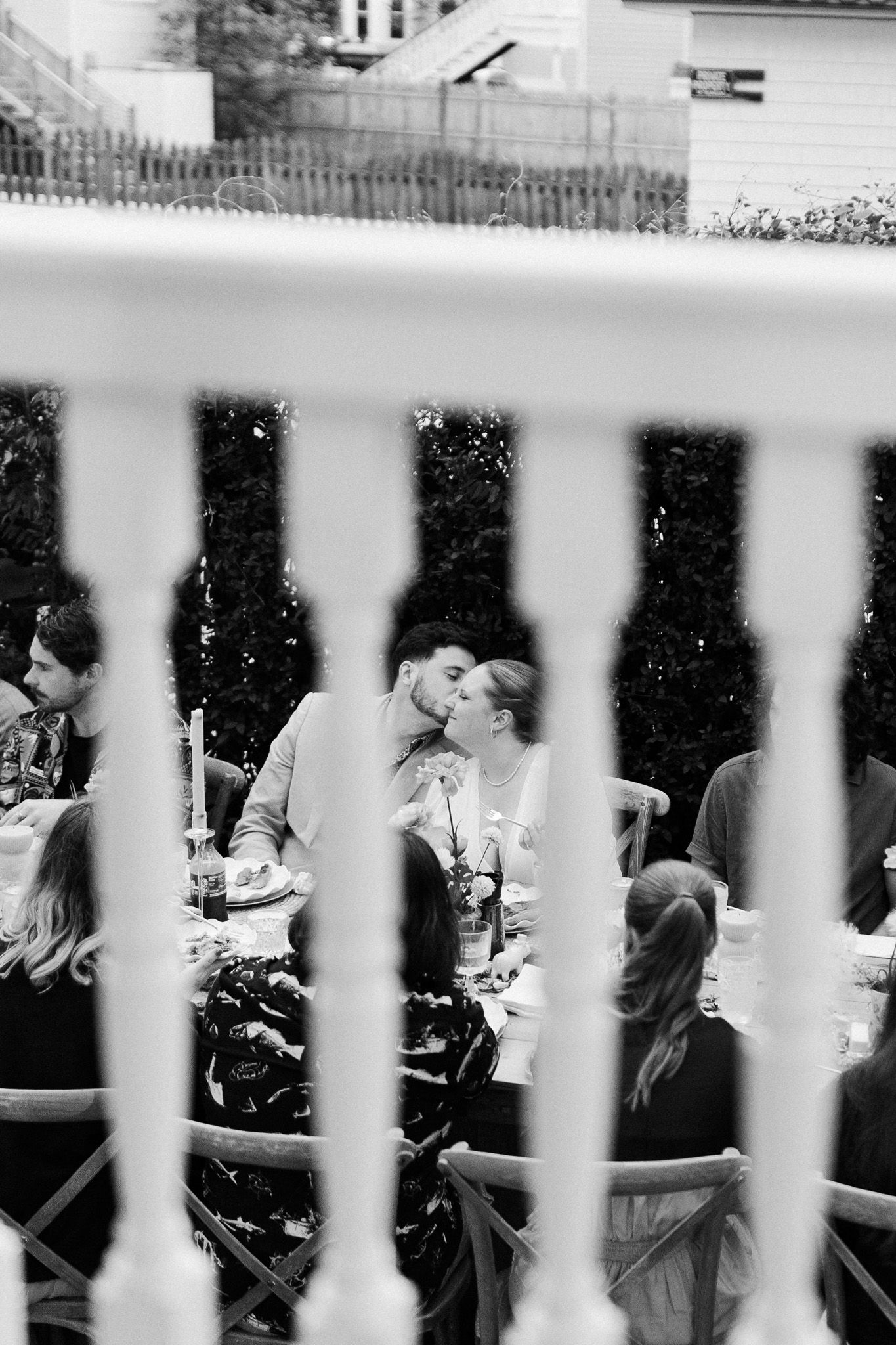 Black and white photo of a couple sharing a kiss at an outdoor garden dinner party, viewed through white fence rails.