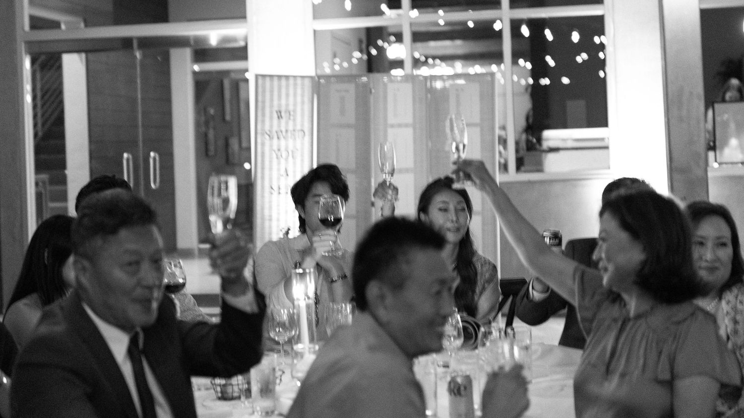 Black and white photo of a group of people raising glasses in a toast at a restaurant celebration dinner.