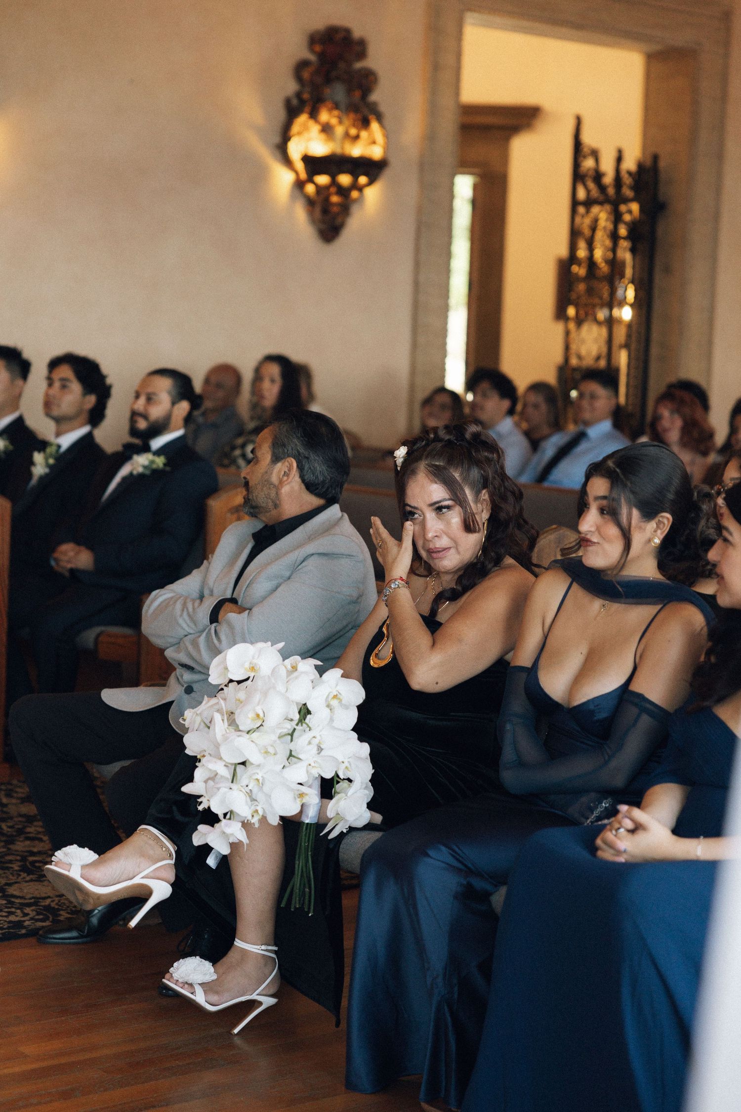Wedding guests in navy blue dresses hold white floral bouquets while seated in an elegant venue during ceremony.