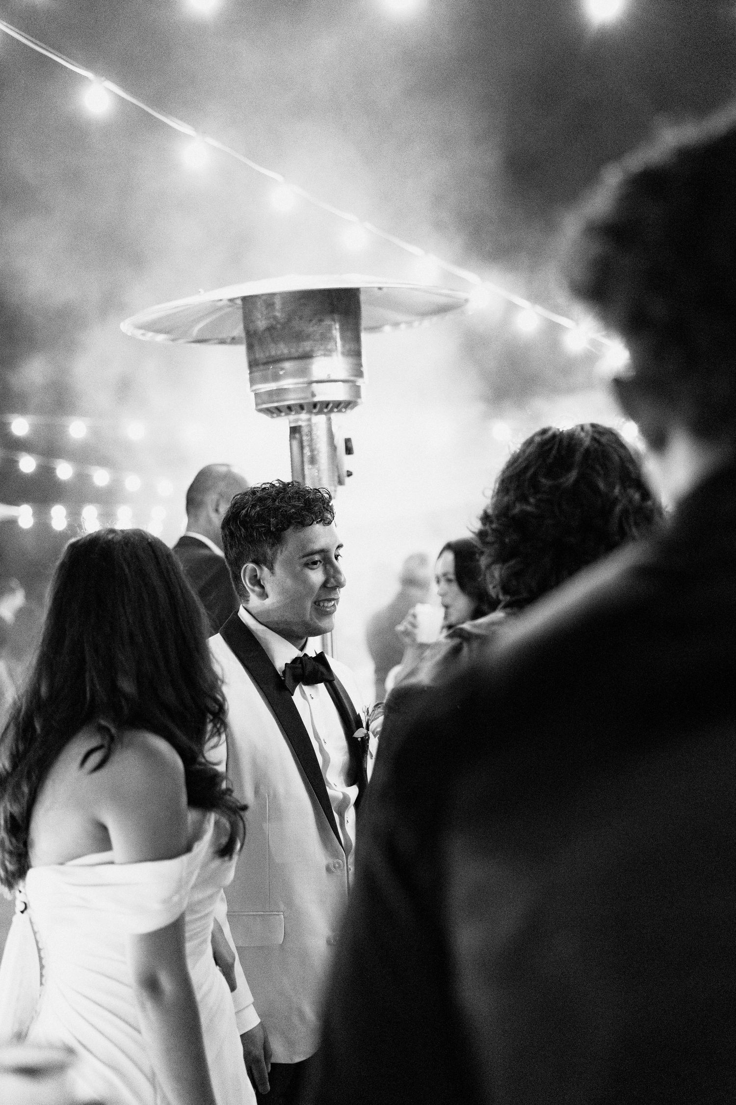 Black and white photo of an outdoor evening event with string lights and a patio heater illuminating the gathering.
