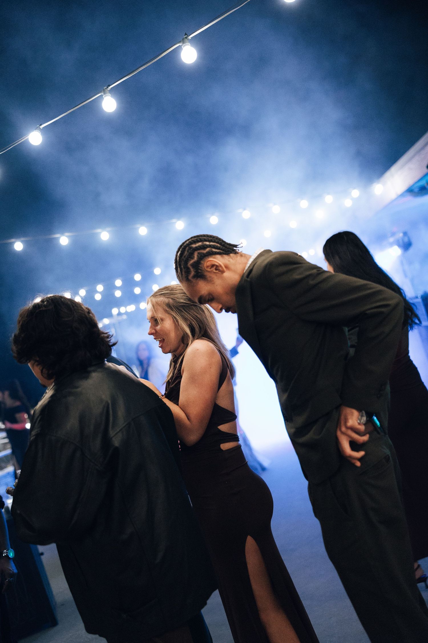 Silhouetted crowd dancing under string lights at a nighttime party with atmospheric blue lighting and fog effects.