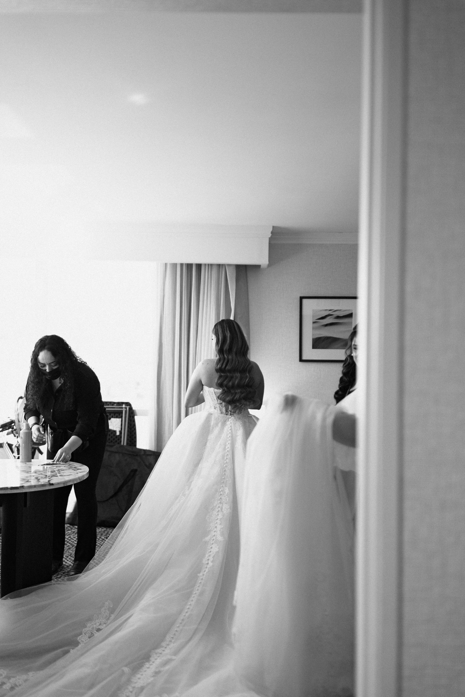 Black and white photo of a bride in a flowing wedding gown being assisted by a helper in an elegant hotel room.