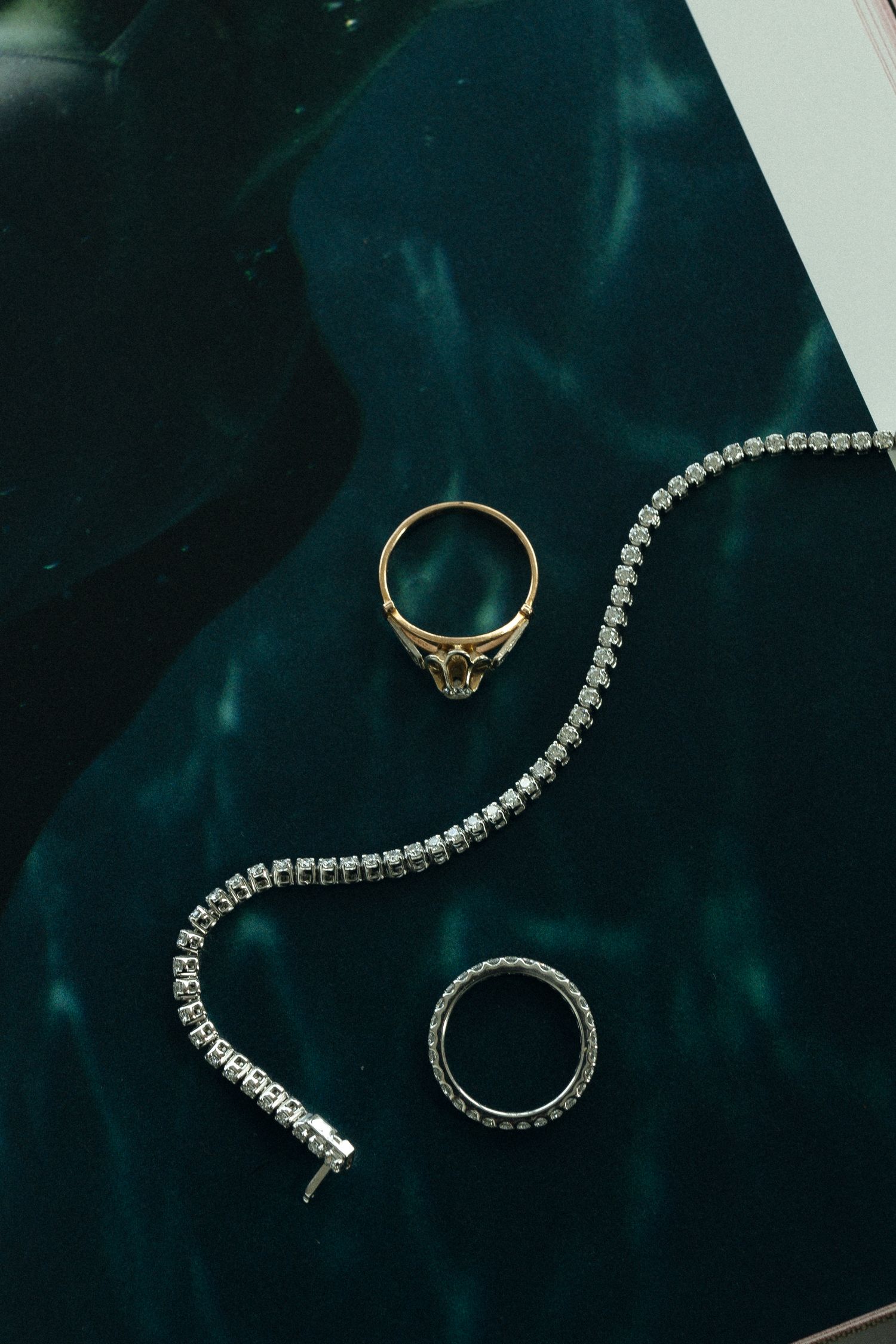 Two elegant rings and a delicate tennis bracelet with diamonds displayed on a deep teal velvet surface.
