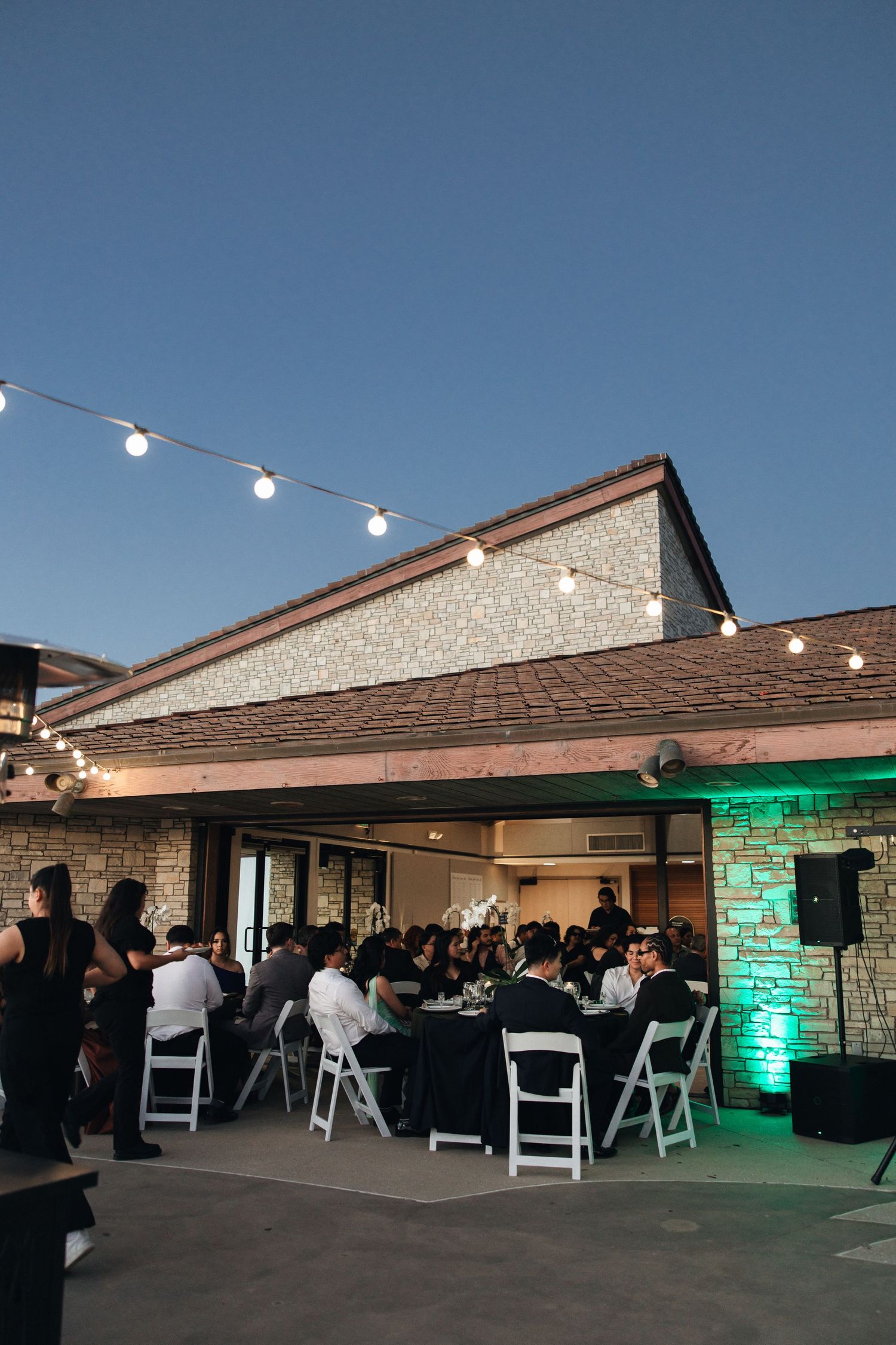 Evening outdoor reception with string lights and green uplighting at a stone venue with guests seated at tables.