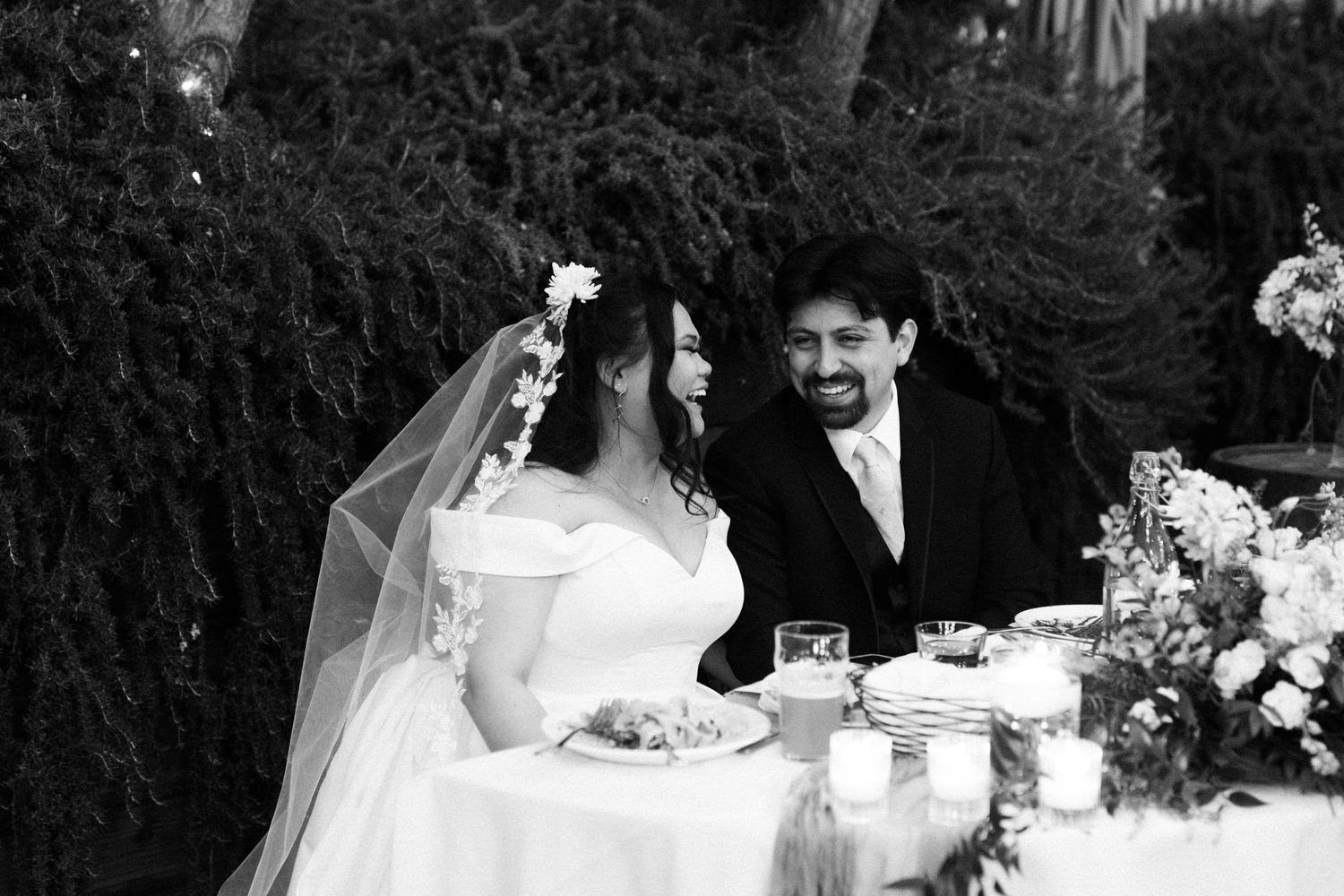 A vintage black and white photograph shows a couple celebrating at an outdoor wedding reception. Documentary/ photojournalist photography