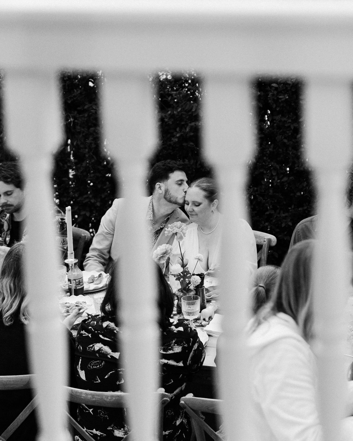 Black and white photo of a couple sharing a kiss at an outdoor wedding reception dinner table.