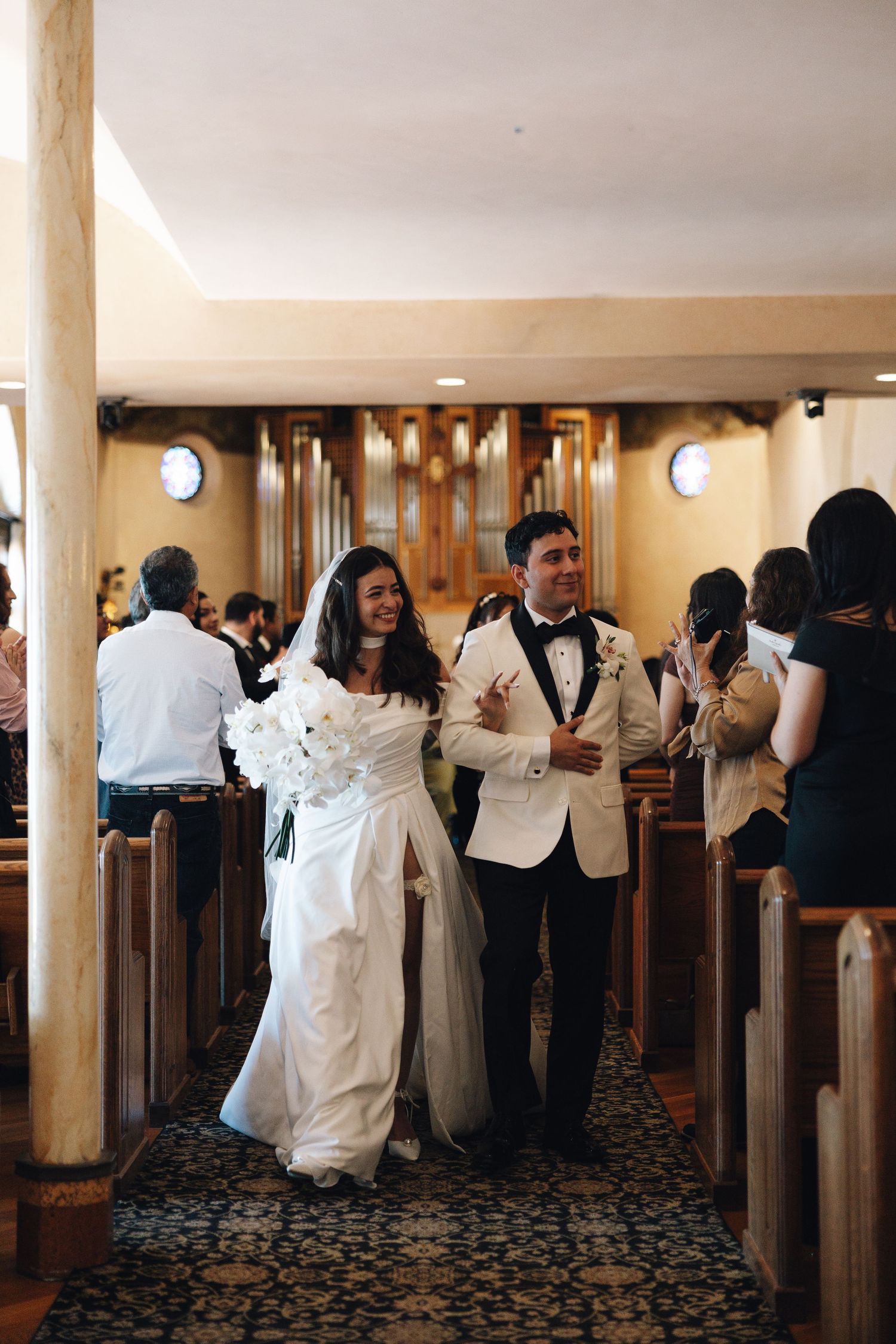 Wedding guests in formal black and white attire socialize in an elegant hallway with carpeted floors and columns.