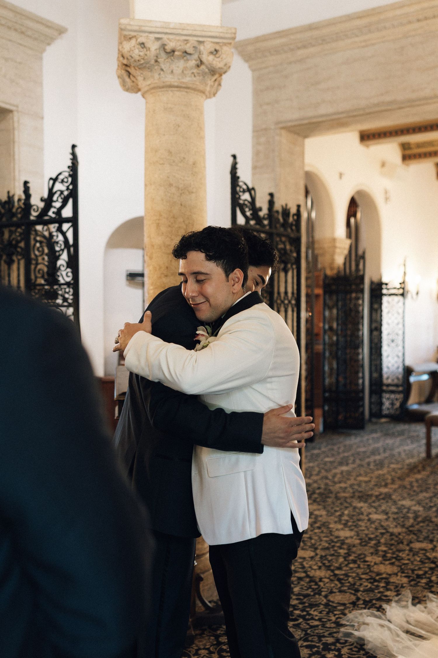 Two people share a warm embrace in an elegant hallway with ornate columns and wrought iron details.